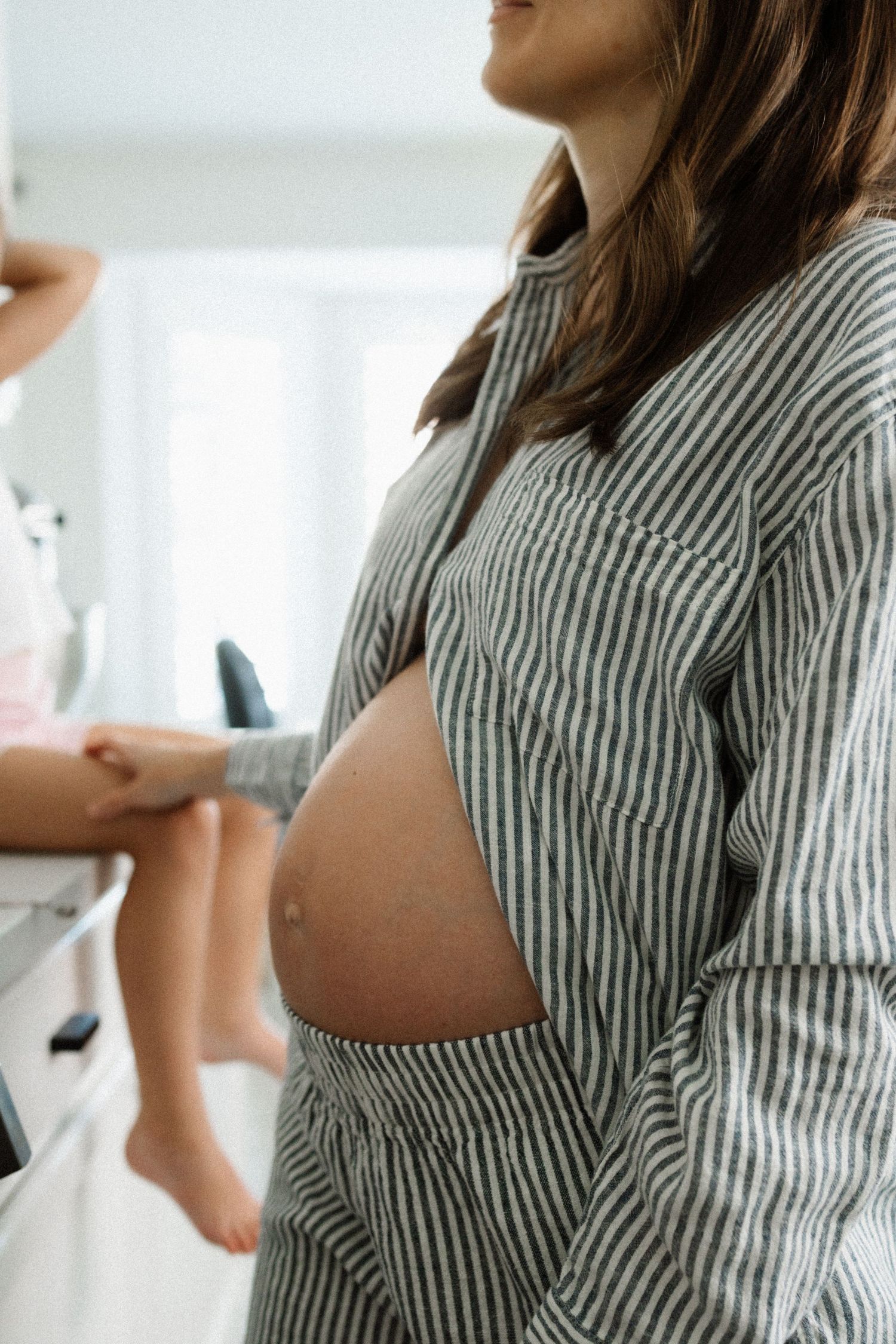 Side view of pregnant woman in striped shirt showing bare belly in bright indoor setting with soft natural light.