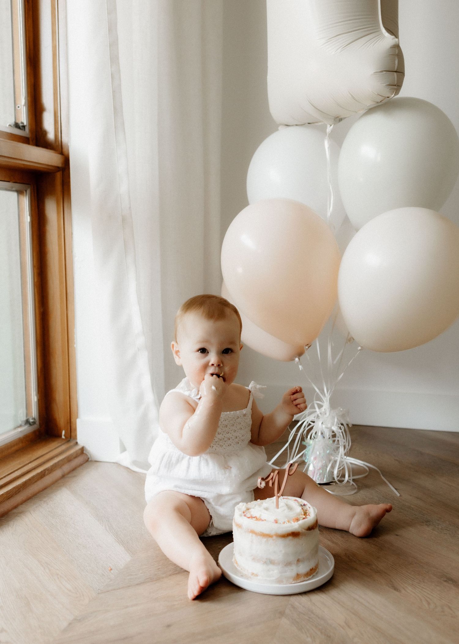 Baby in white dress sits beside birthday cake and neutral balloons by sunny window.