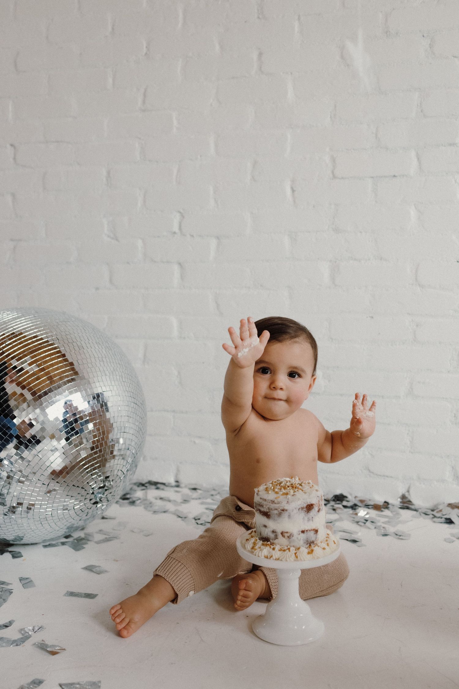 Baby reaching up with both hands near disco ball and white cake against brick wall.