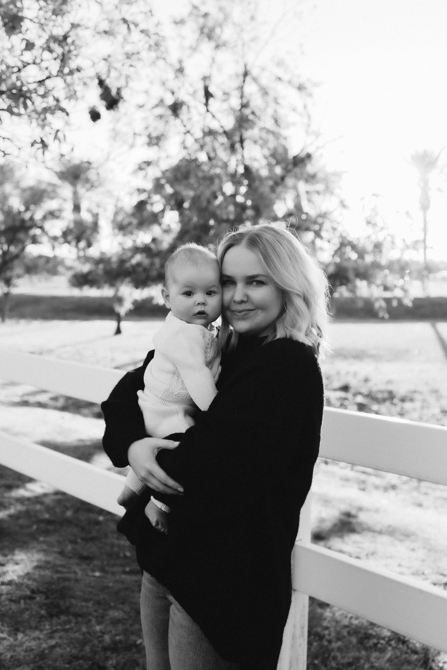 Mother and baby pose together near a white fence in black and white photography.