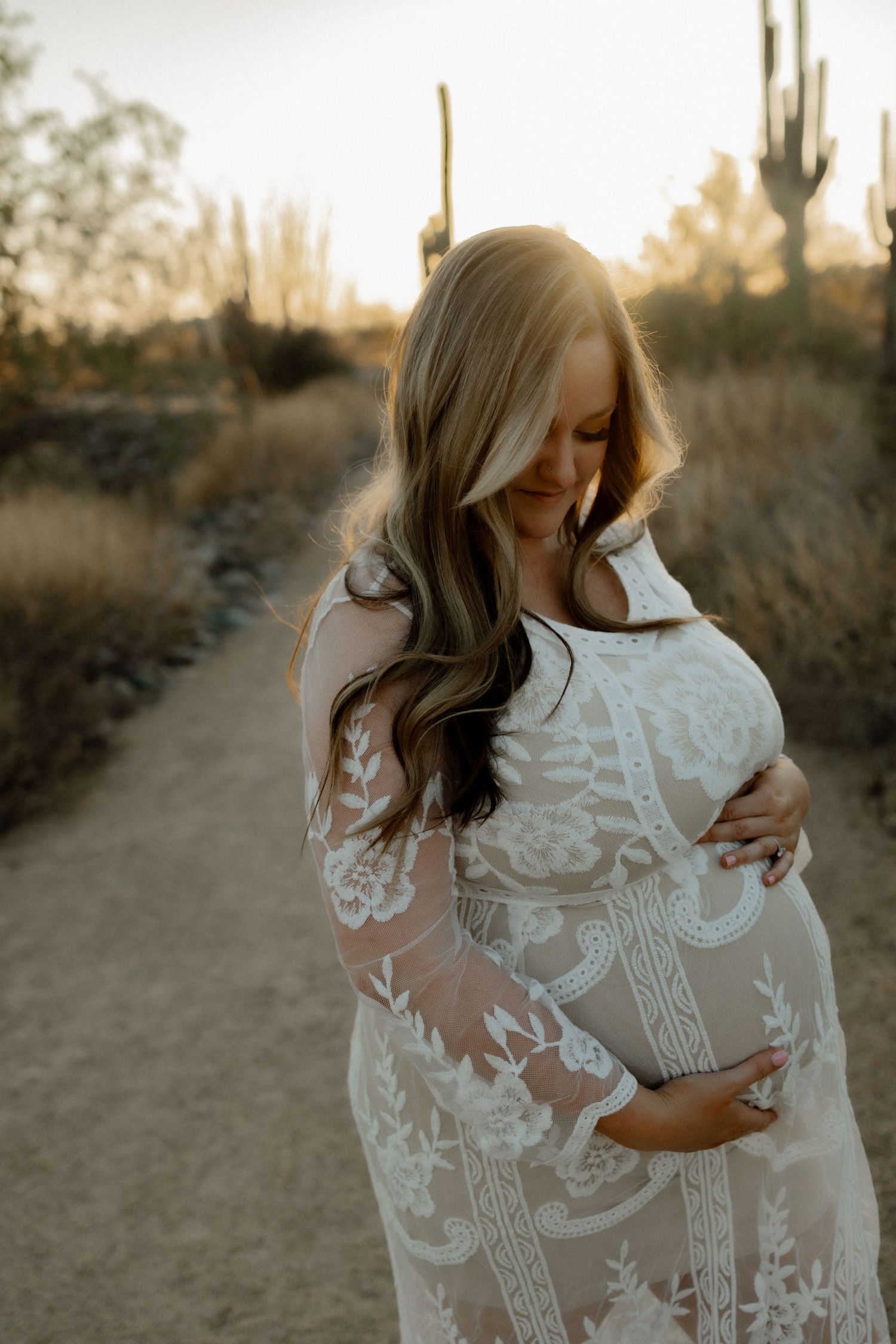 A pregnant woman in a white lace dress poses in a desert setting during golden hour.