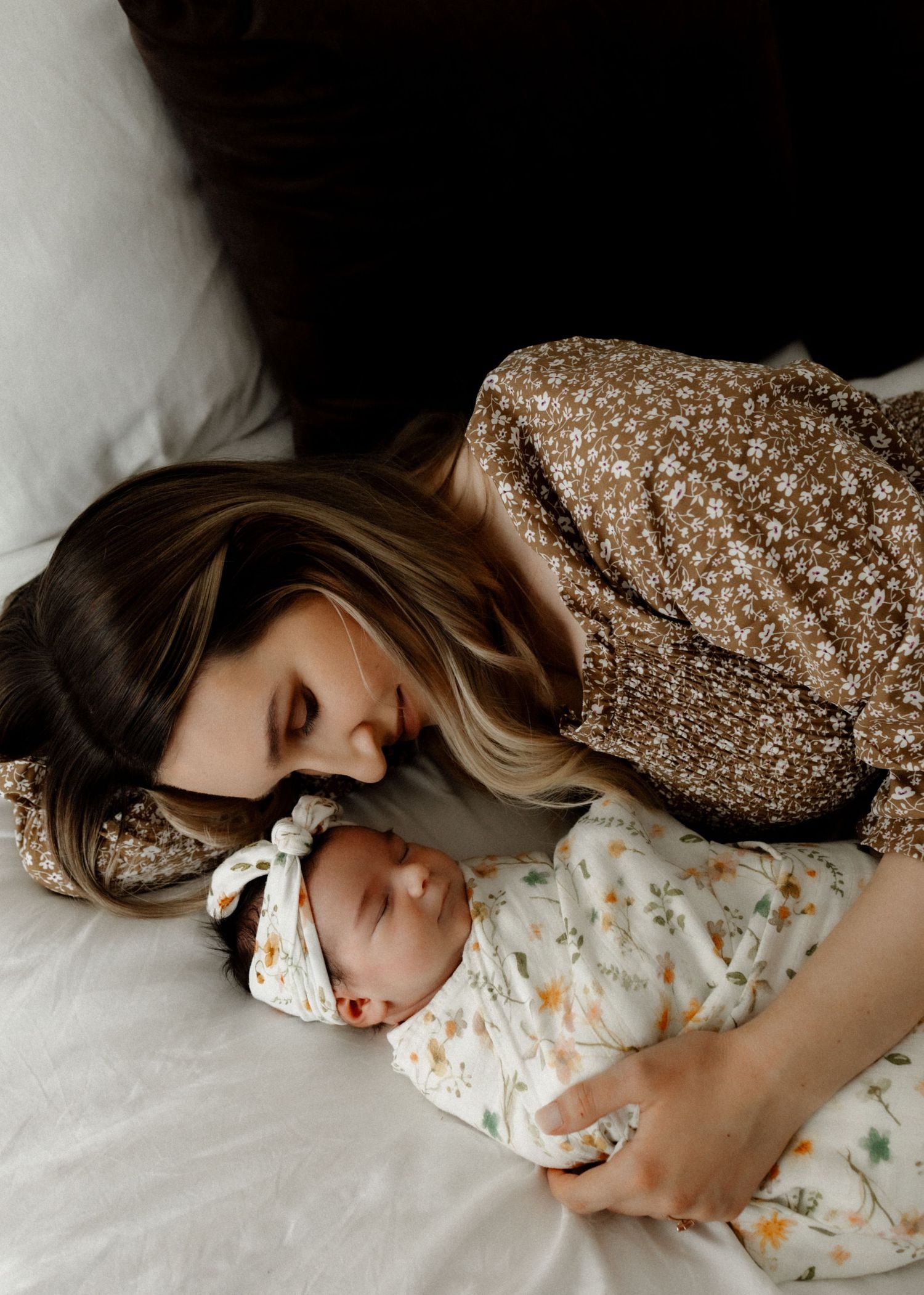 Parent in floral dress lying with sleeping newborn on white bedding.