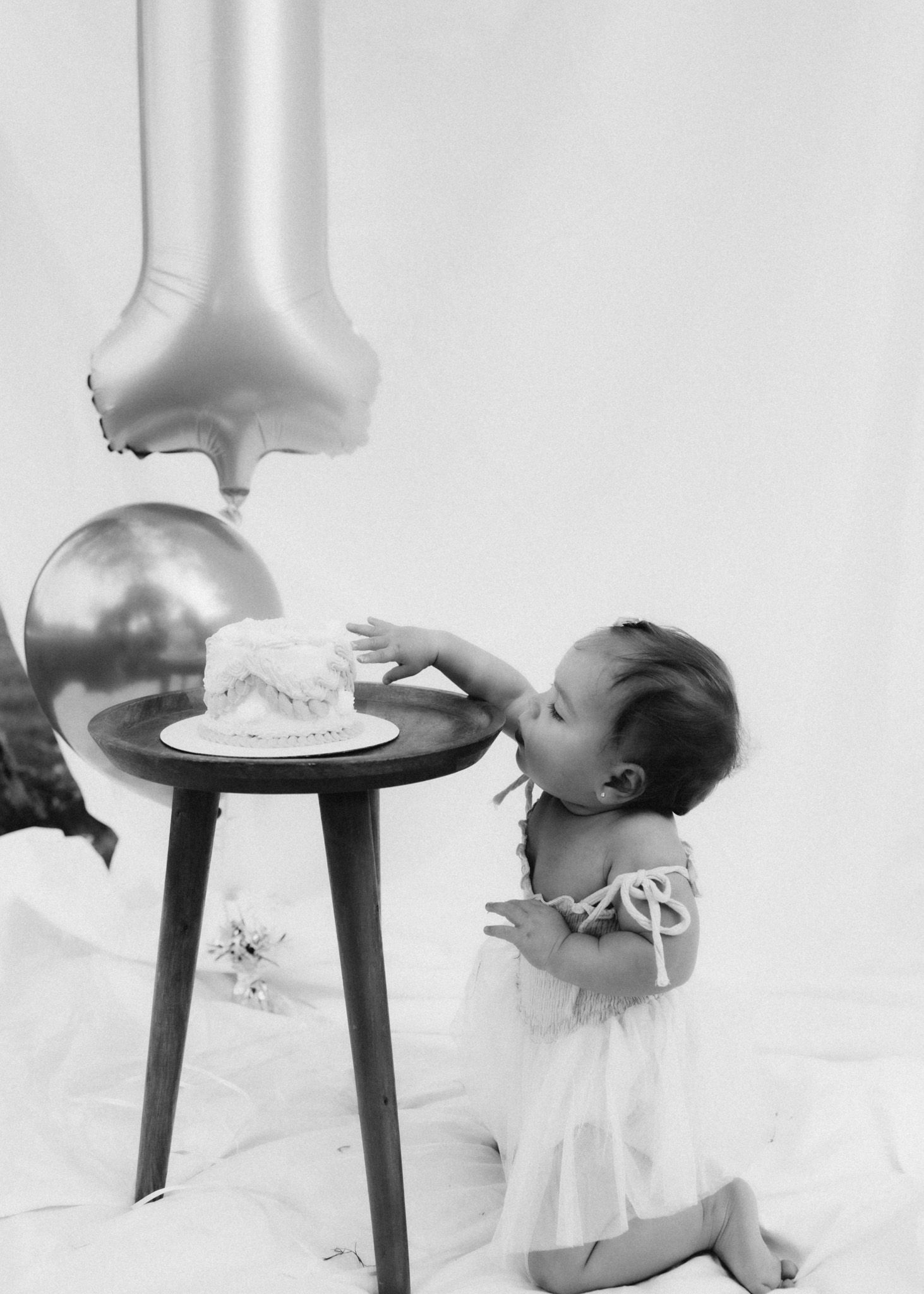 A baby in a white dress reaching for a small cake on a table with balloons in a minimalist setting.