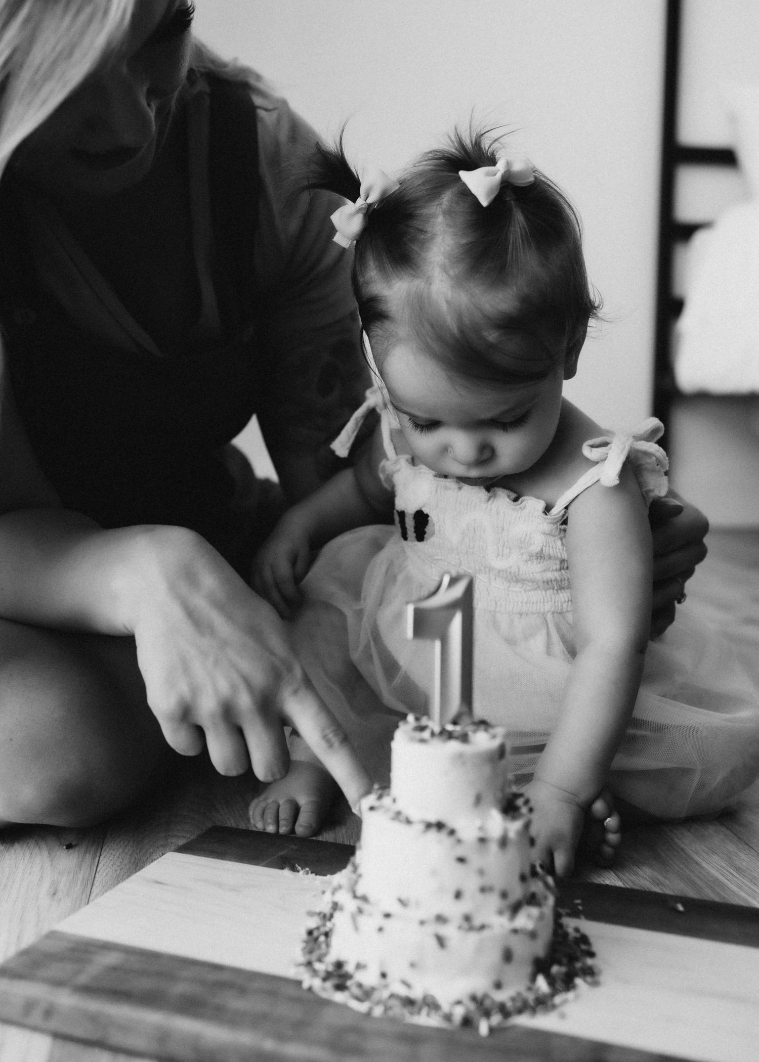 Black and white photo of toddler in white dress looking at first birthday cake on wooden board.