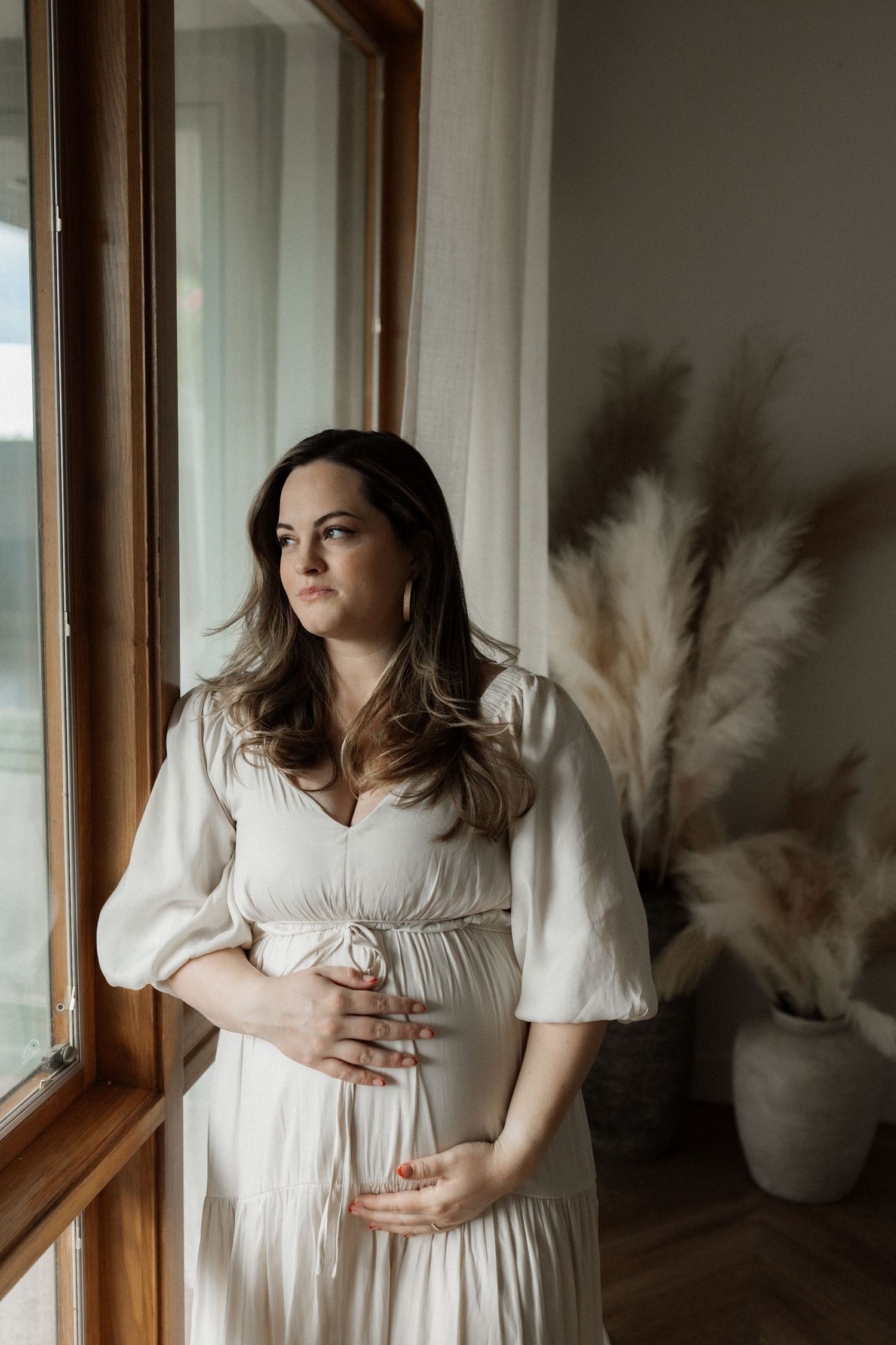 A figure in a flowing white dress with puff sleeves stands by a window with pampas grass decor in a moody setting.
