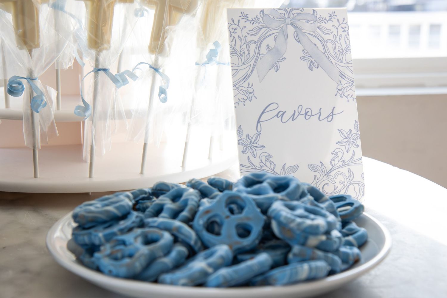 Blue chocolate-covered pretzels on a white plate beside a elegant favors sign at a baby shower.