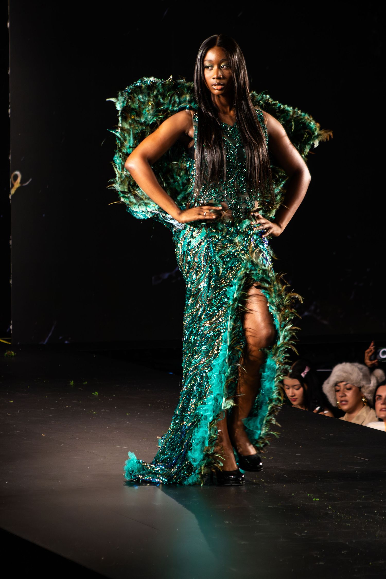 Model walks runway in stunning emerald green sequined evening gown with high slit during fashion show performance.