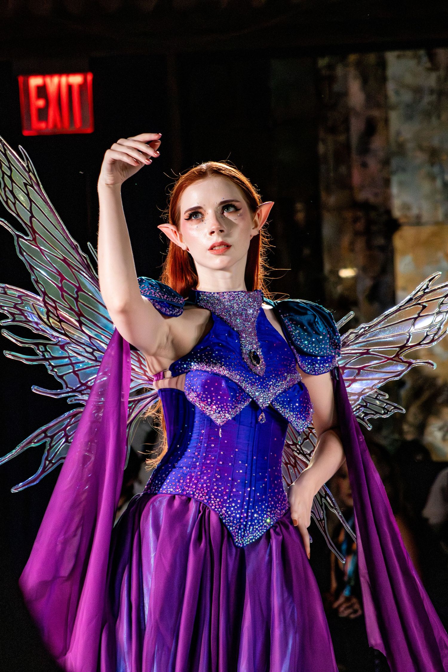 Model wears purple fantasy costume with iridescent wings and crystal embellishments.