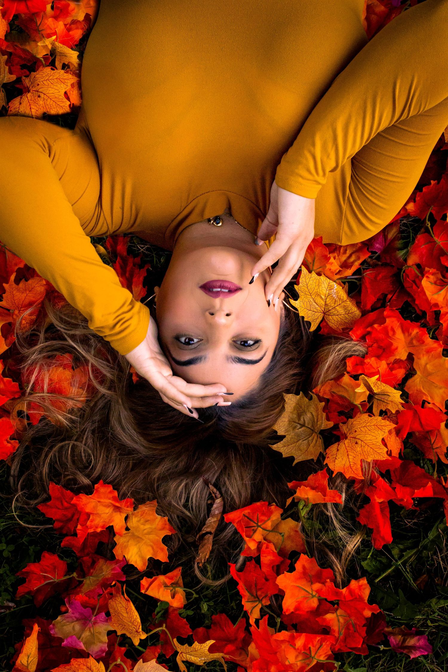 Portrait lying on vibrant autumn leaves wearing mustard yellow clothing against red foliage background.