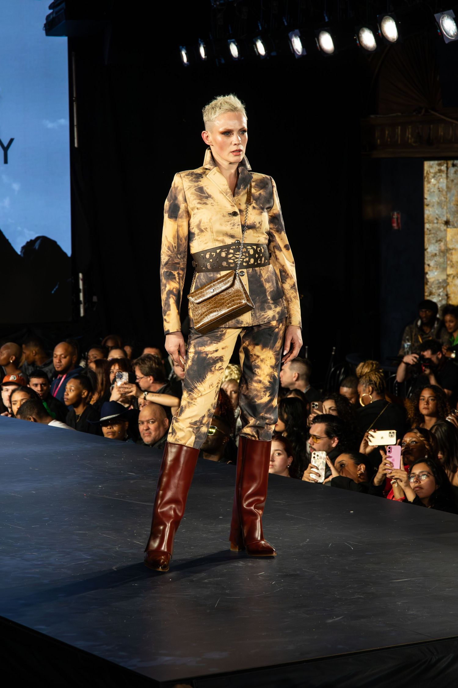 A model walks the runway wearing a brown distressed military-inspired outfit with tall leather boots at a fashion show.