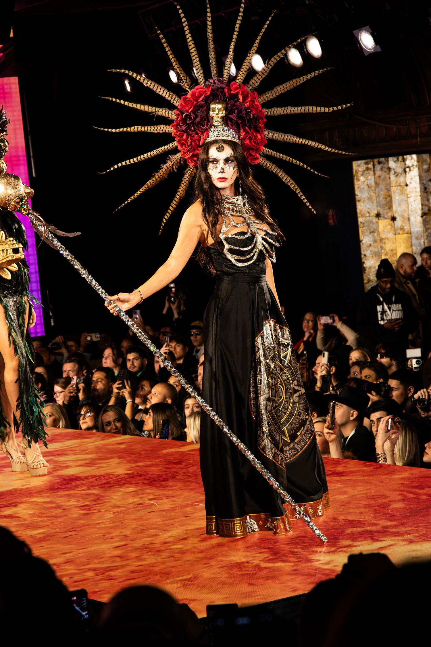 A performer in elaborate Dia de los Muertos costume with feathered headdress walks runway during cultural fashion show.