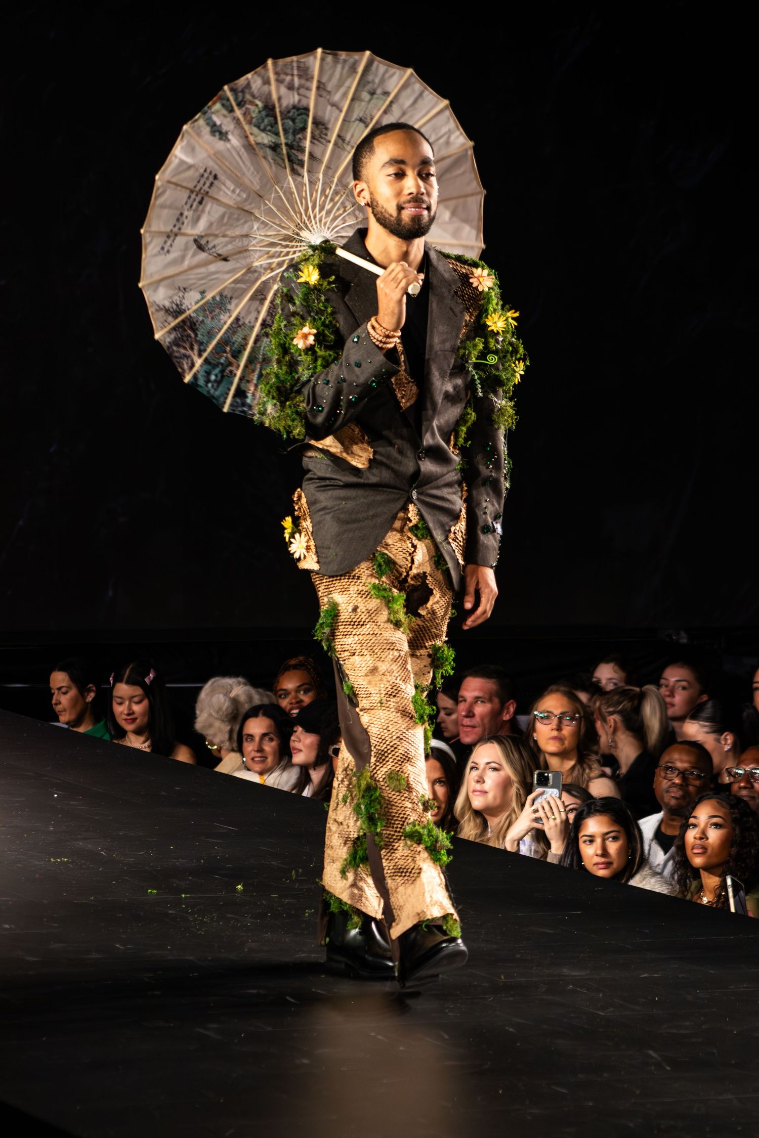 A model walks the runway wearing a dark shirt, floral-accented camouflage pants, and holding a decorative parasol.