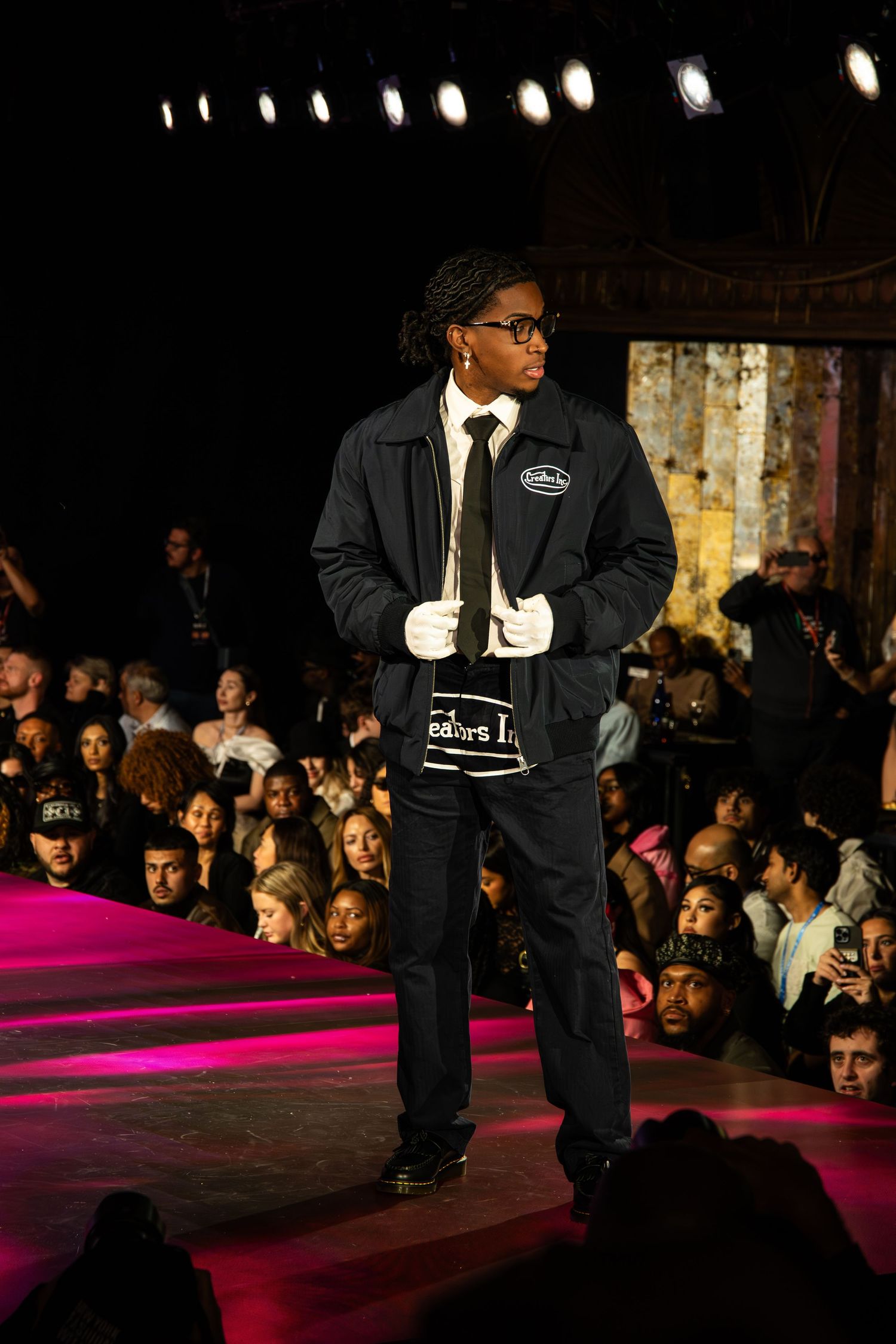 A model walks the runway at a fashion show wearing a dark suit with white gloves on a pink carpet stage.