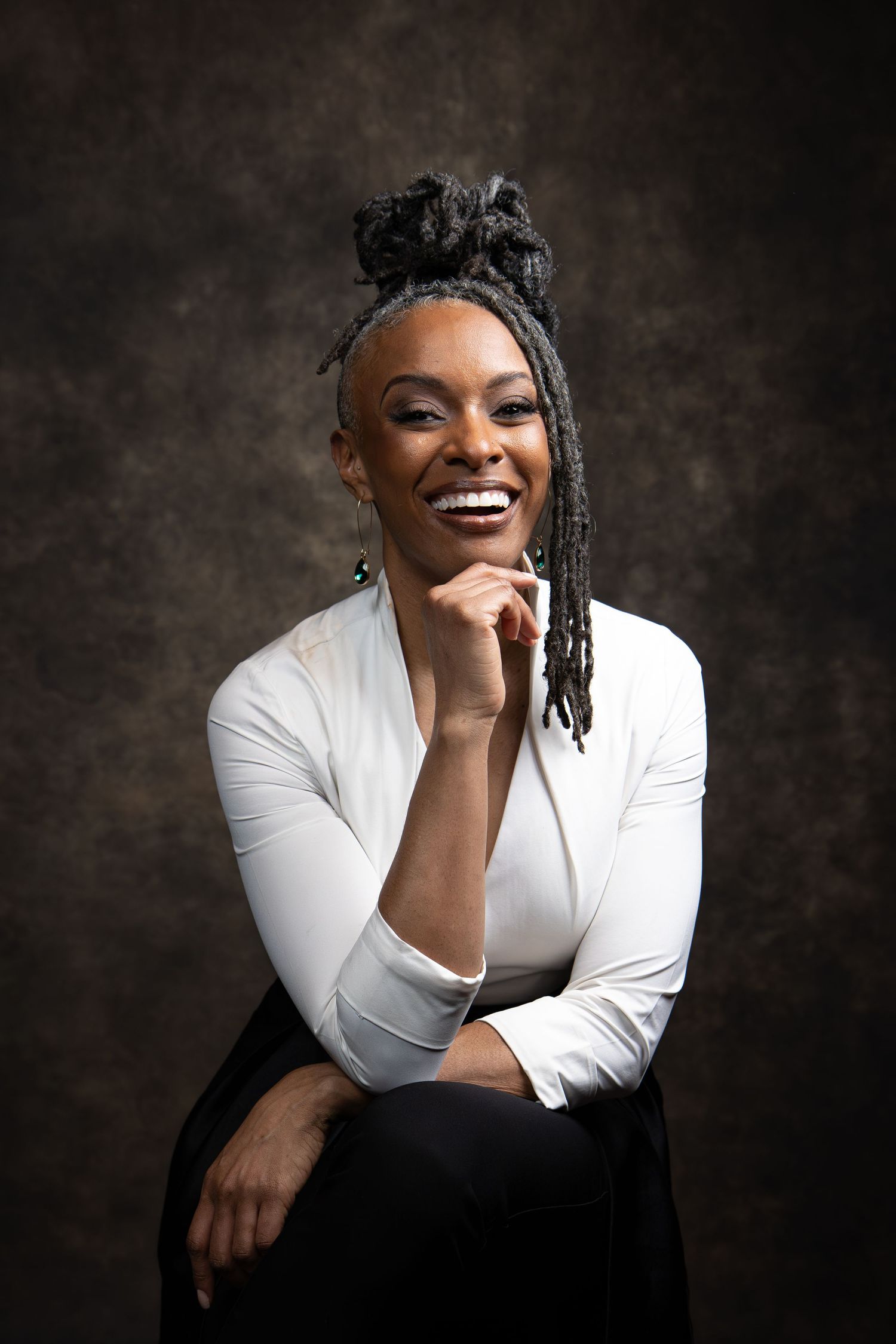 Smiling Black woman with locs updo, wearing white top, posing confidently against dark studio background.
