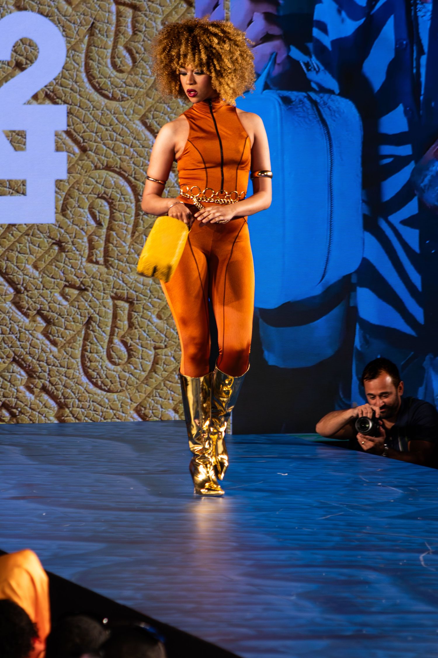 Female model walks runway in rust orange bodysuit with gold boots and yellow clutch at fashion show.