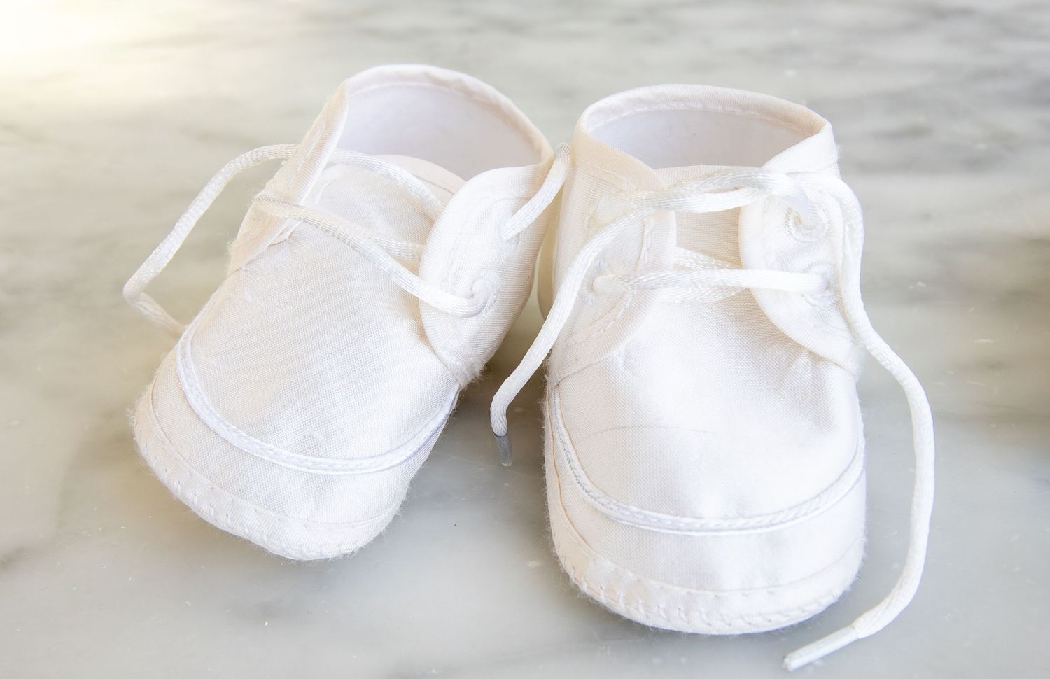 White baby shoes with ribbon laces on marble surface, perfect for christening or special occasions.