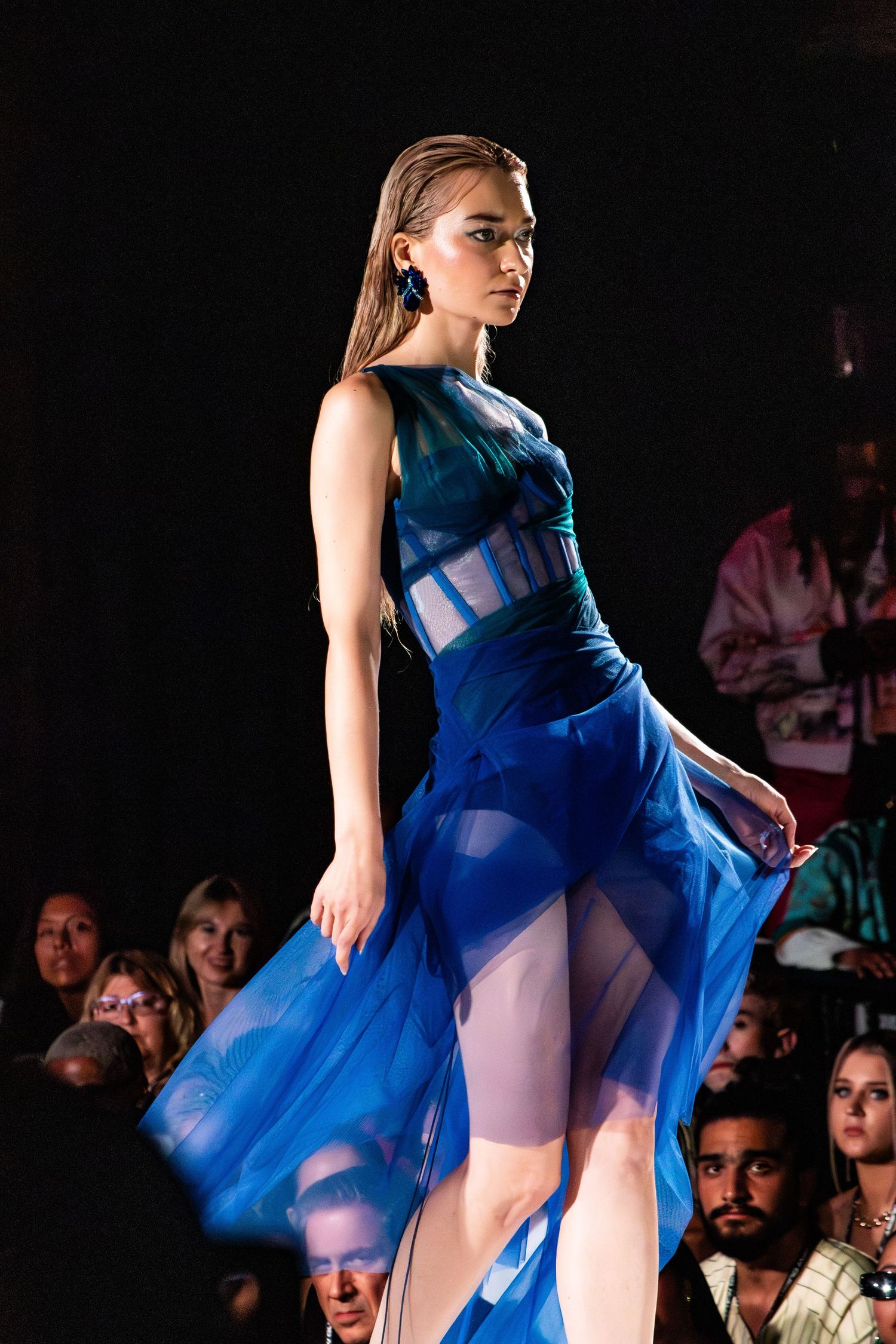 Model walks runway in sheer blue corset dress with tulle overlay.