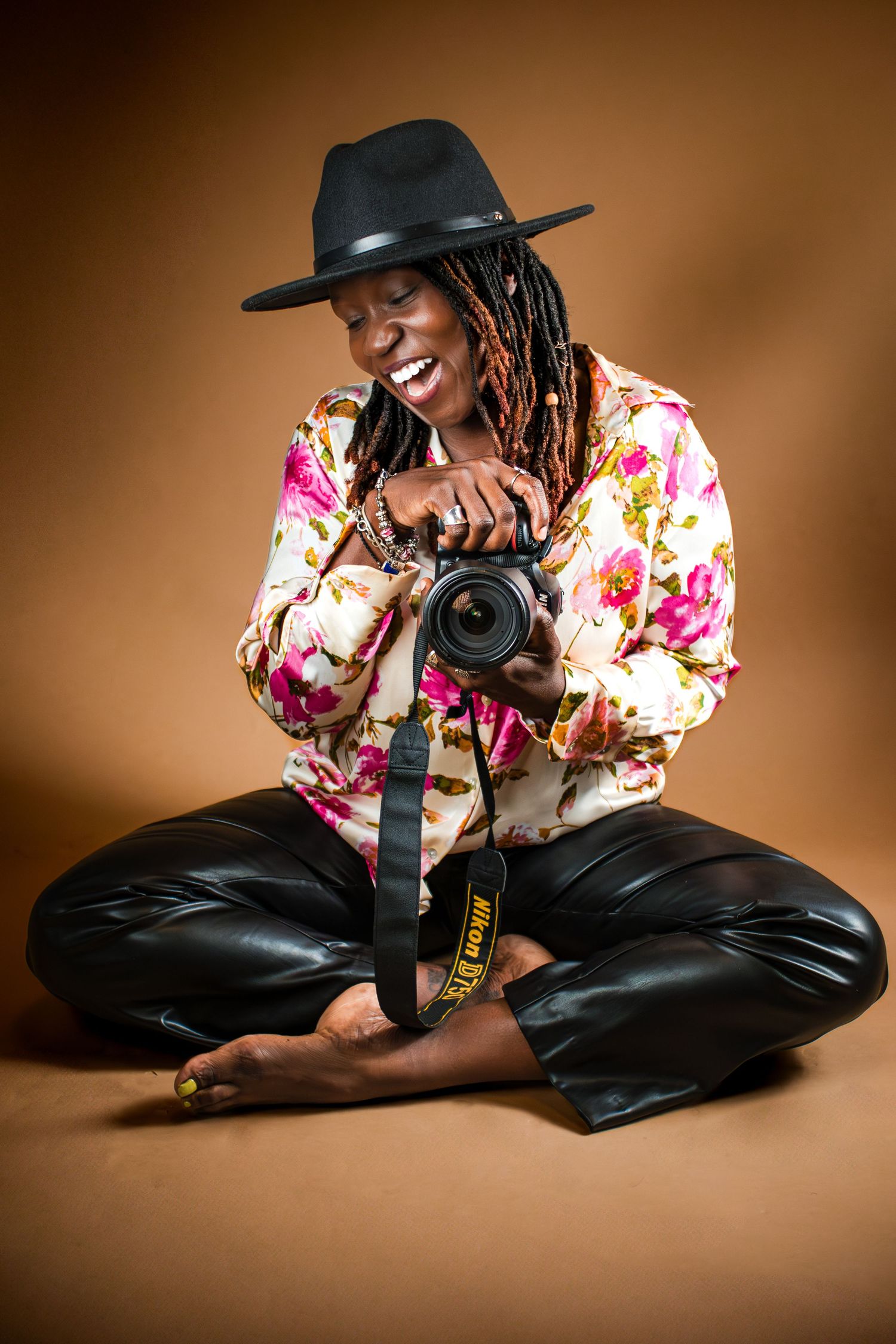 Professional photographer in floral shirt and black hat sits on floor with camera against brown backdrop.