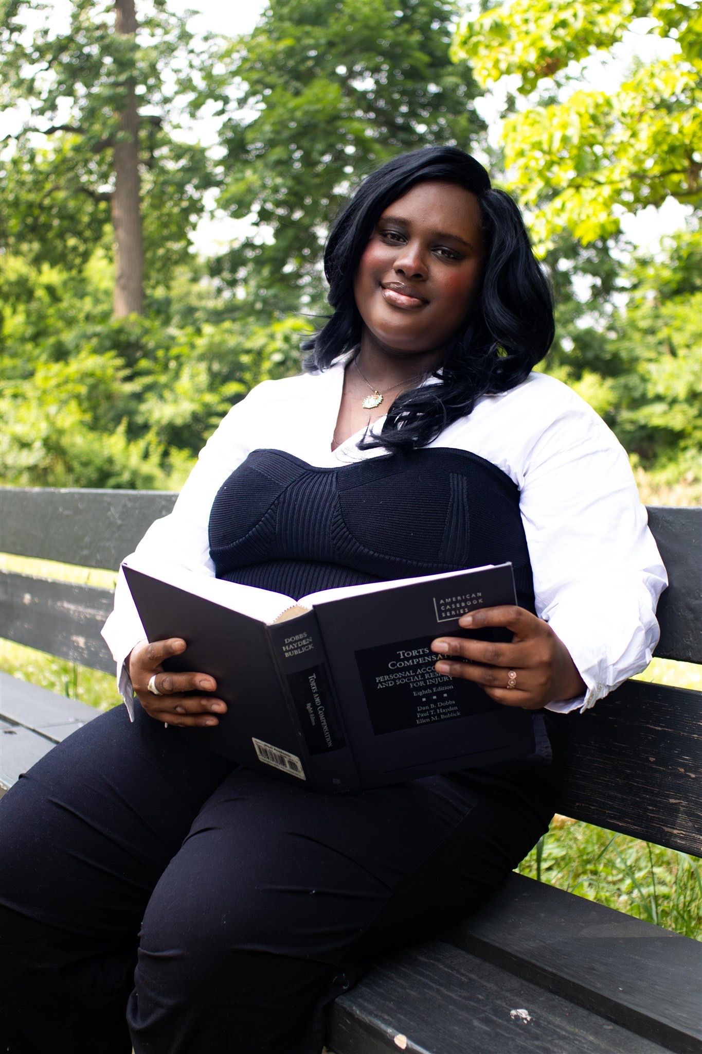 Person reading a book while sitting on a park bench surrounded by lush green trees on a sunny day.