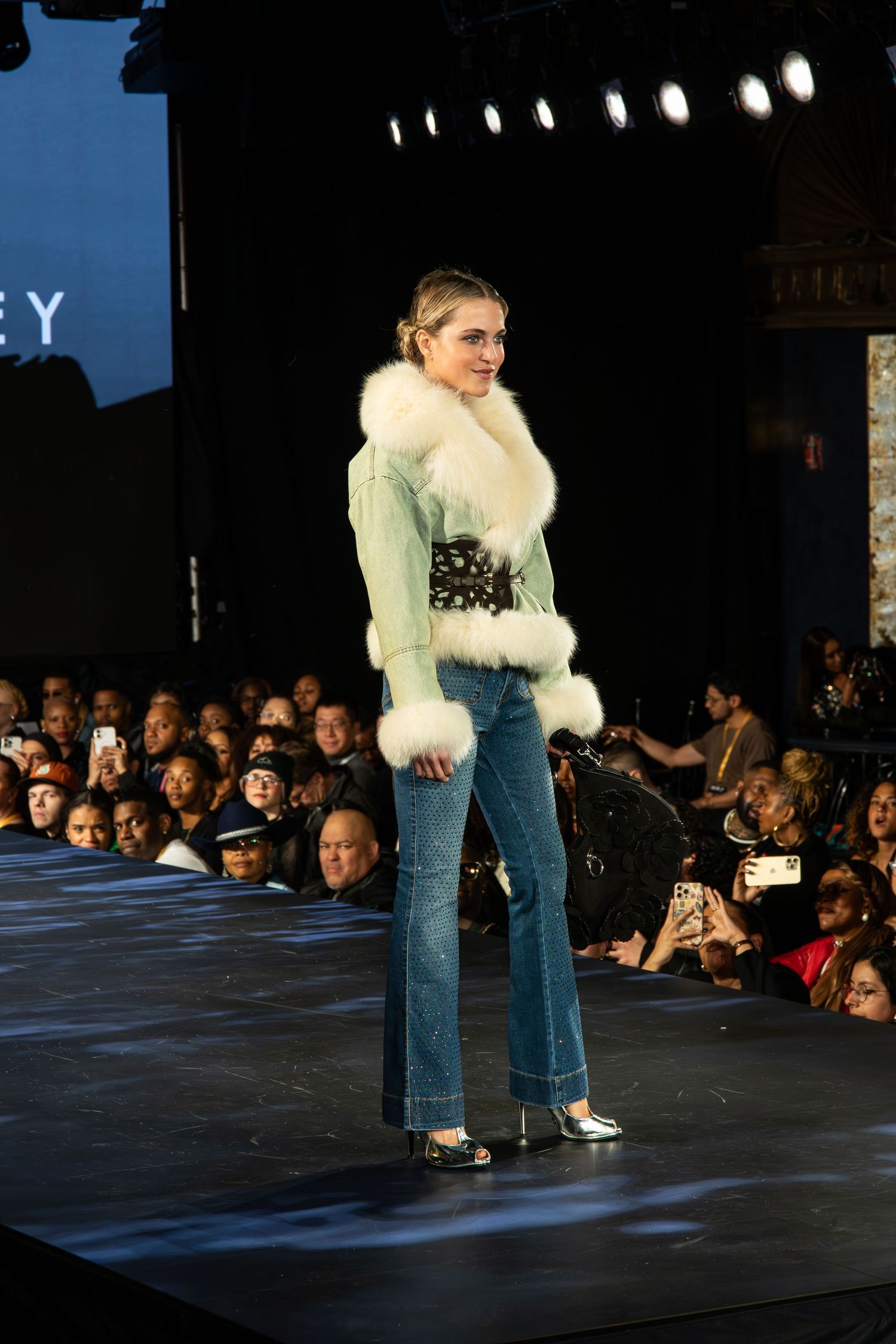 A model walks the runway wearing a mint green fur-trimmed jacket with jeans and silver heels at a fashion show.