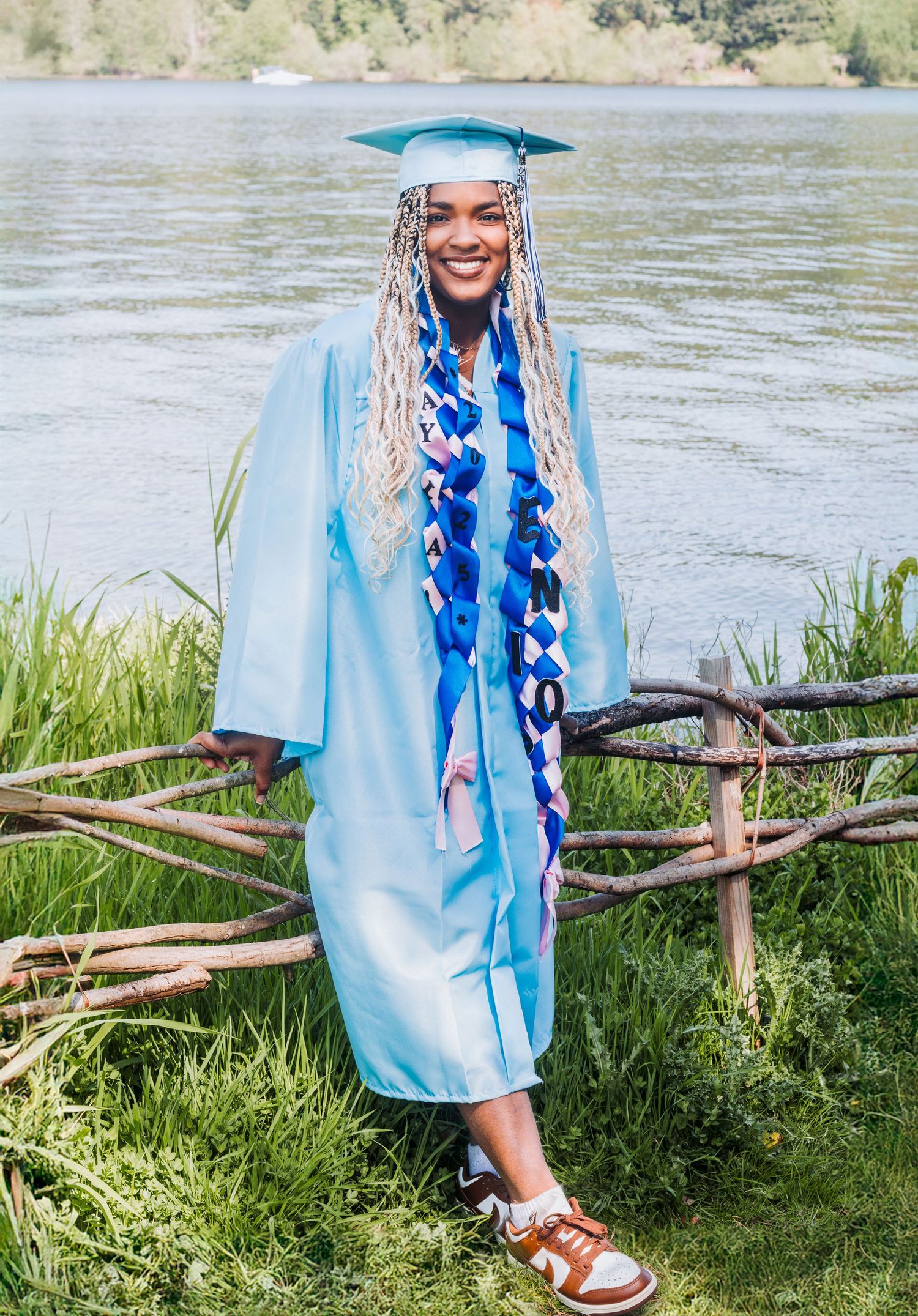 A smiling graduate in a light blue cap and gown with braided hair poses outdoors by a lake.
