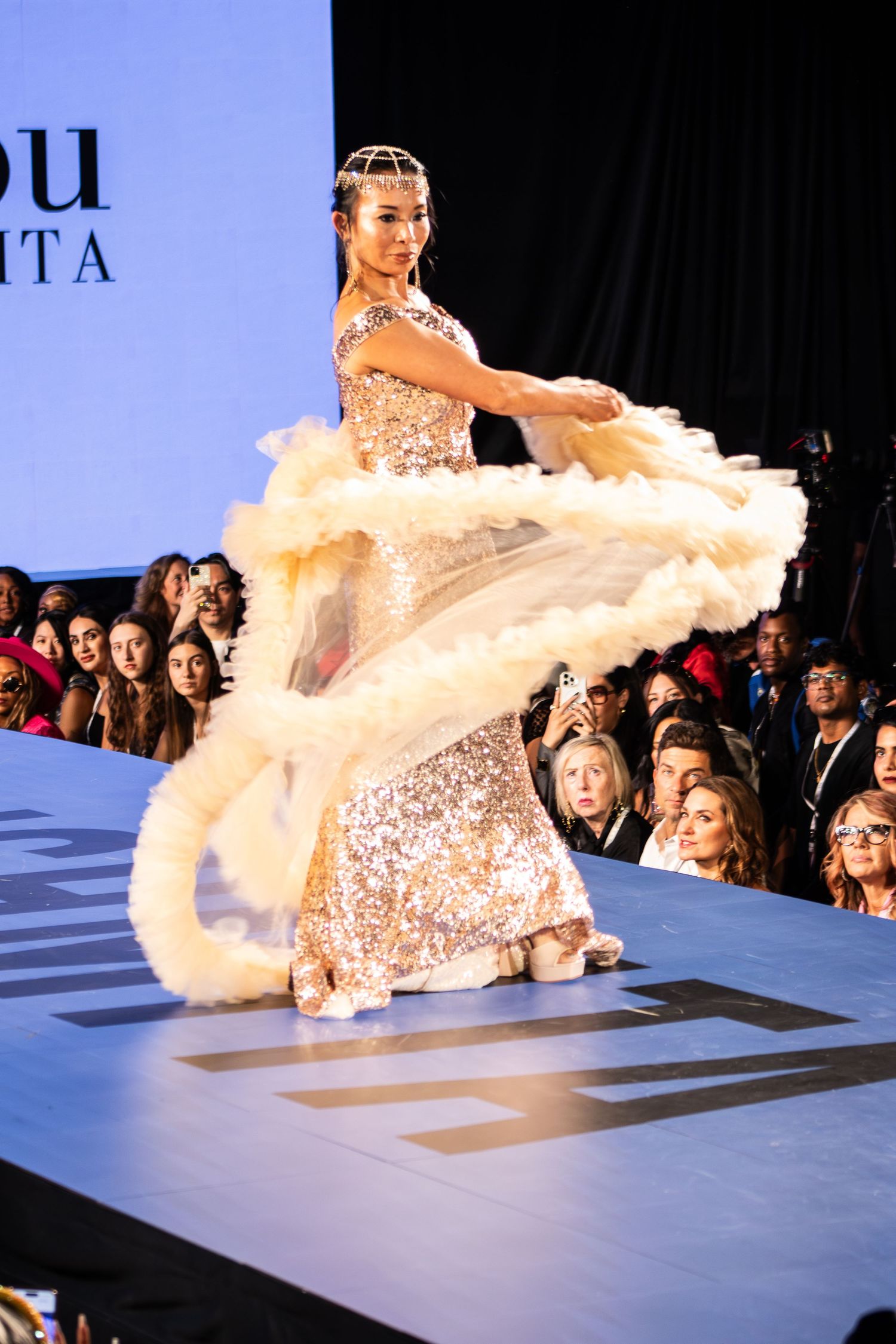 Model in gold sequin gown twirls feather wrap on fashion show runway.