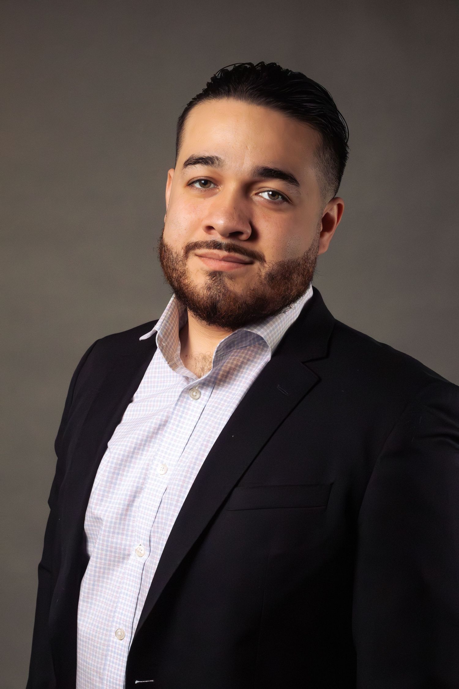Professional headshot of someone in dark suit and checkered shirt against neutral background.