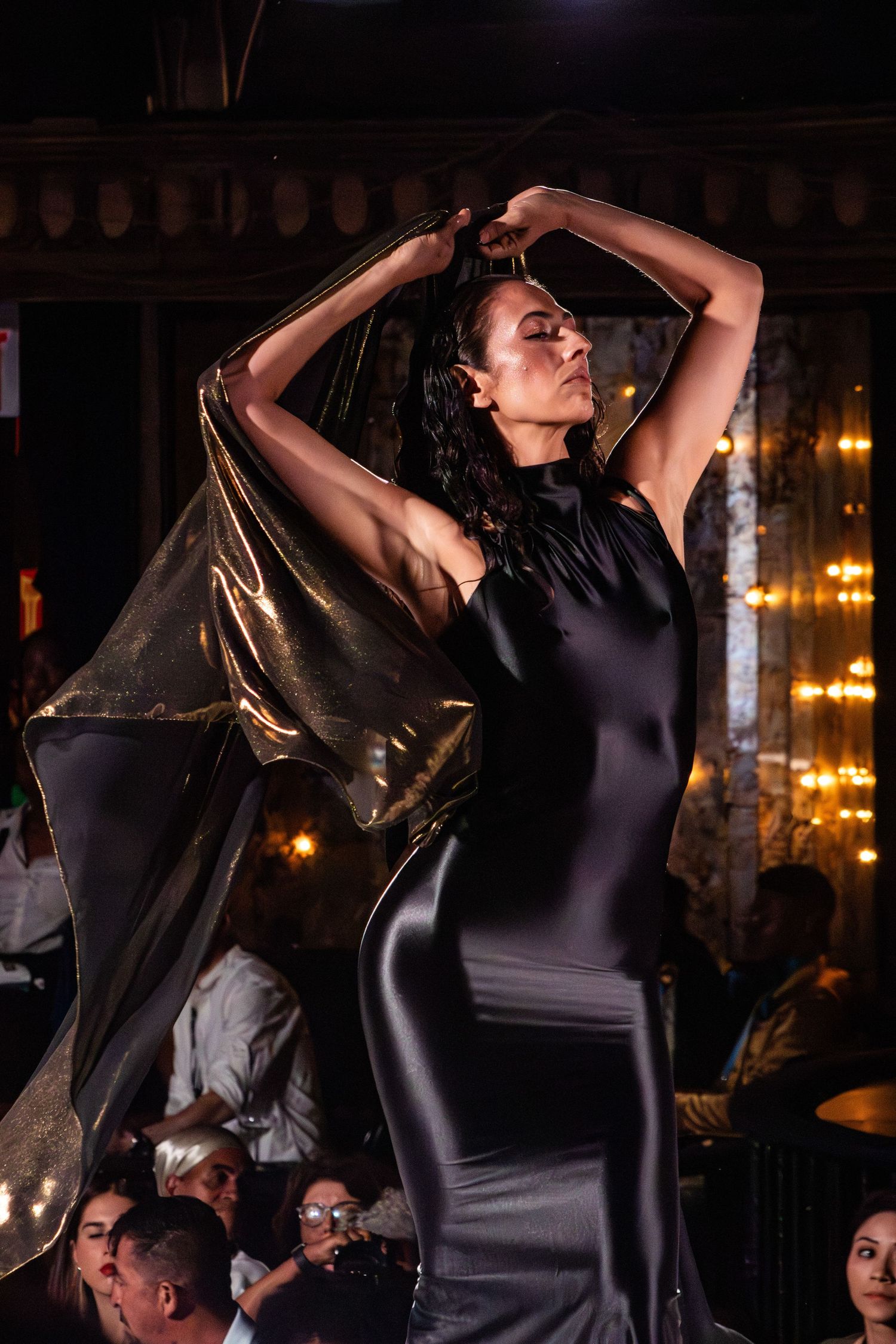 Model in black dress with metallic cape poses dramatically on runway.