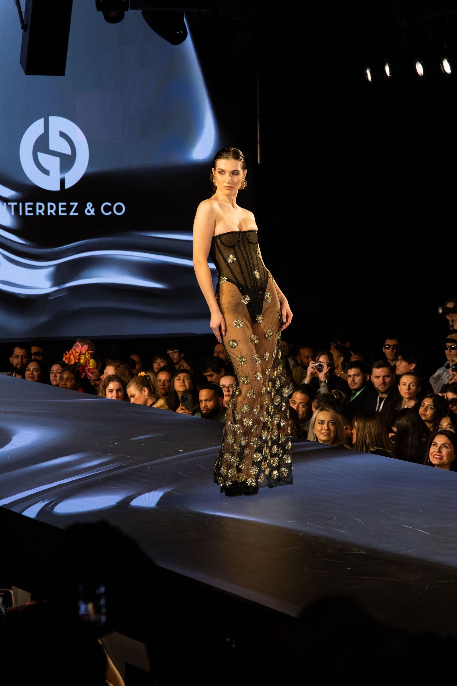 Model walks runway in strapless black evening gown with gold metallic details at fashion show with dramatic lighting.