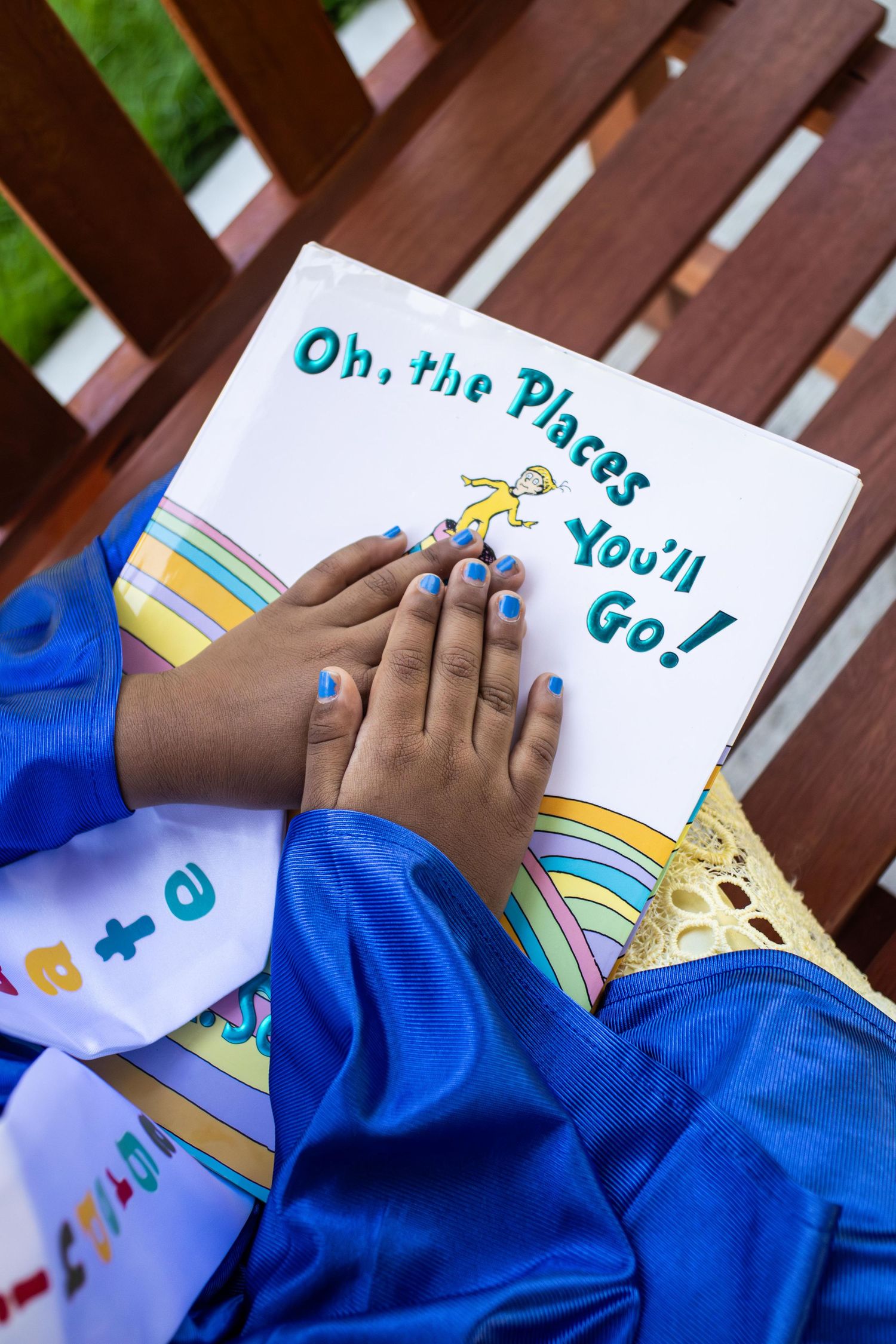 Close-up of hands with blue nail polish holding Dr. Seuss book 'Oh, the Places You'll Go!' while wearing graduation attire.