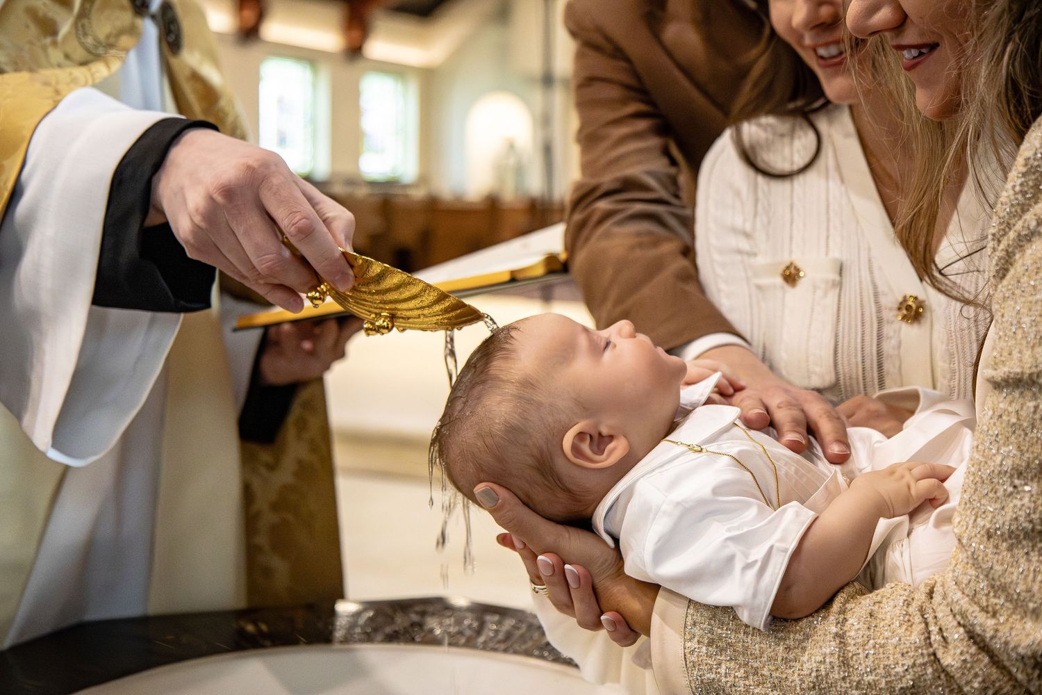 Priest baptizing a newborn baby held by a smiling woman during a Christian baptism ceremony in a church.