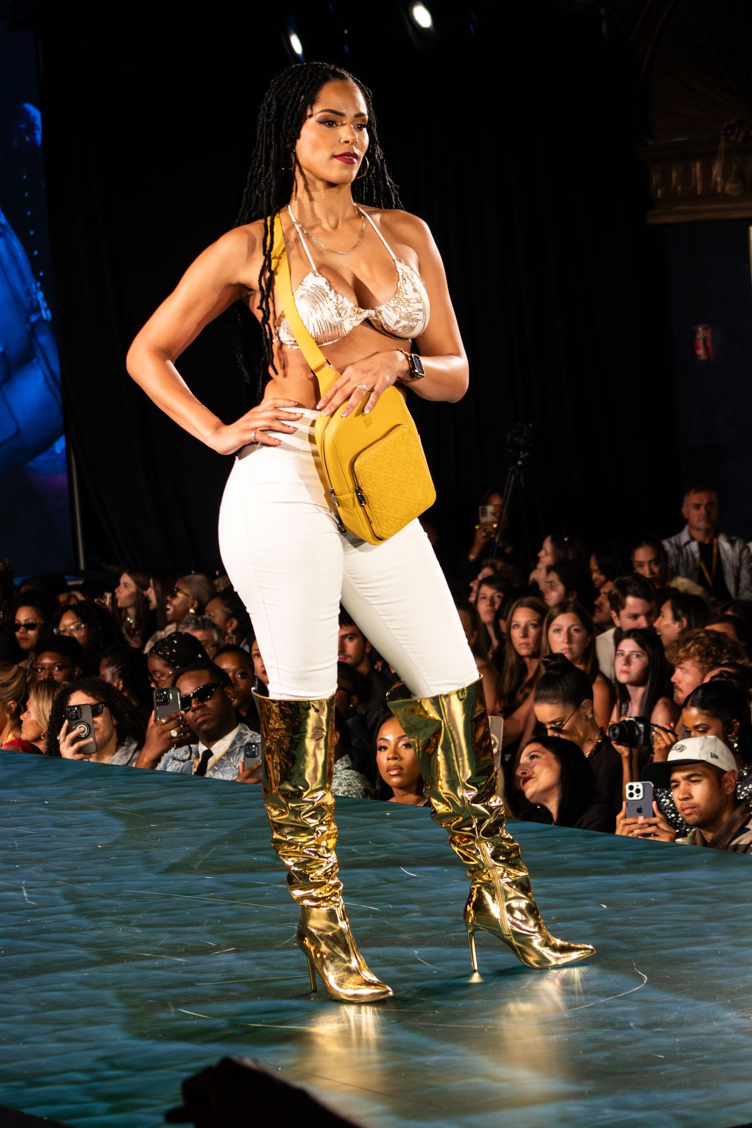 Model walks runway in gold boots, white pants, yellow bag, and lace bralette at a fashion show.