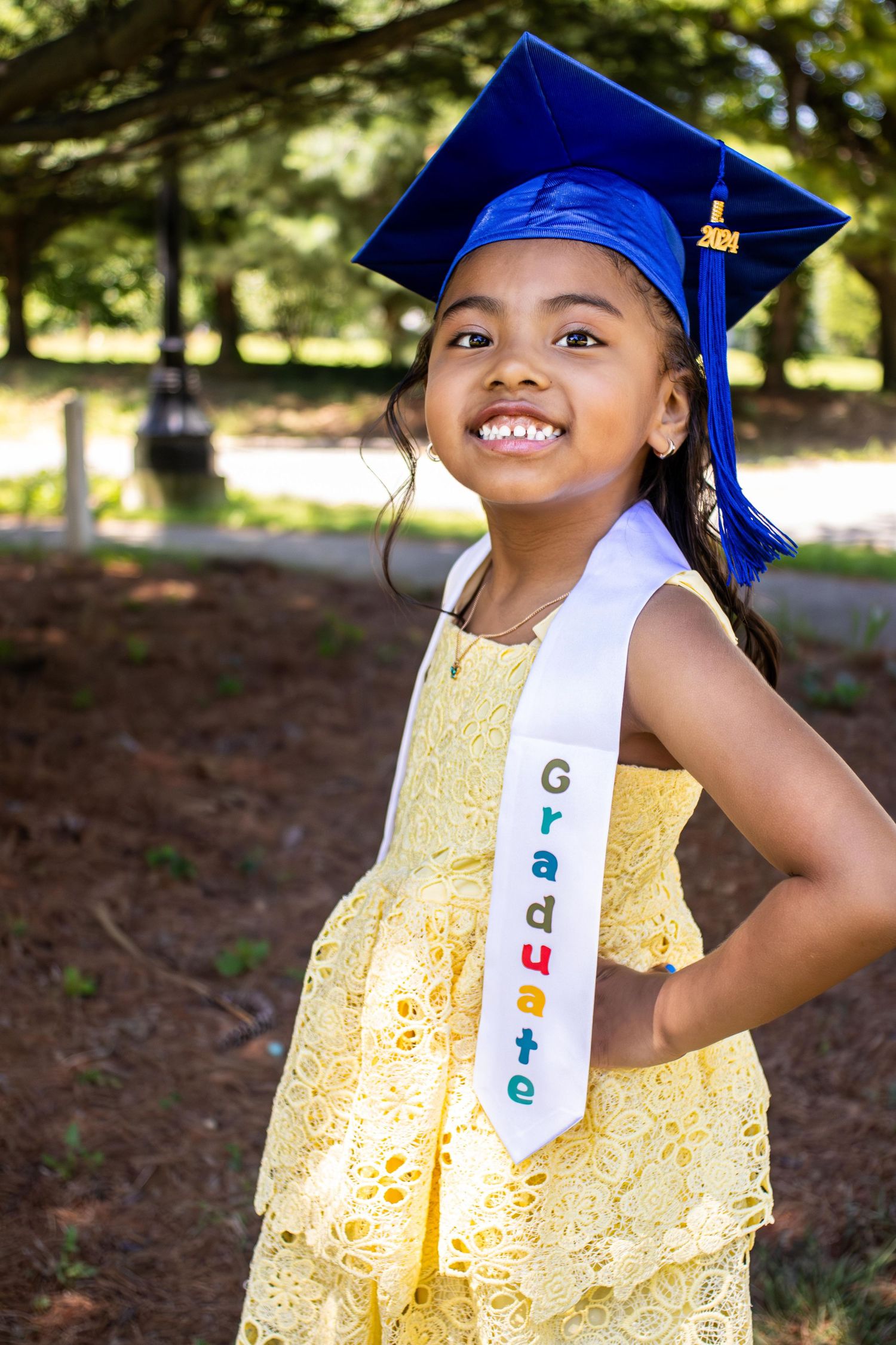 Graduate wearing blue cap and yellow lace dress poses outdoors with a bright smile.