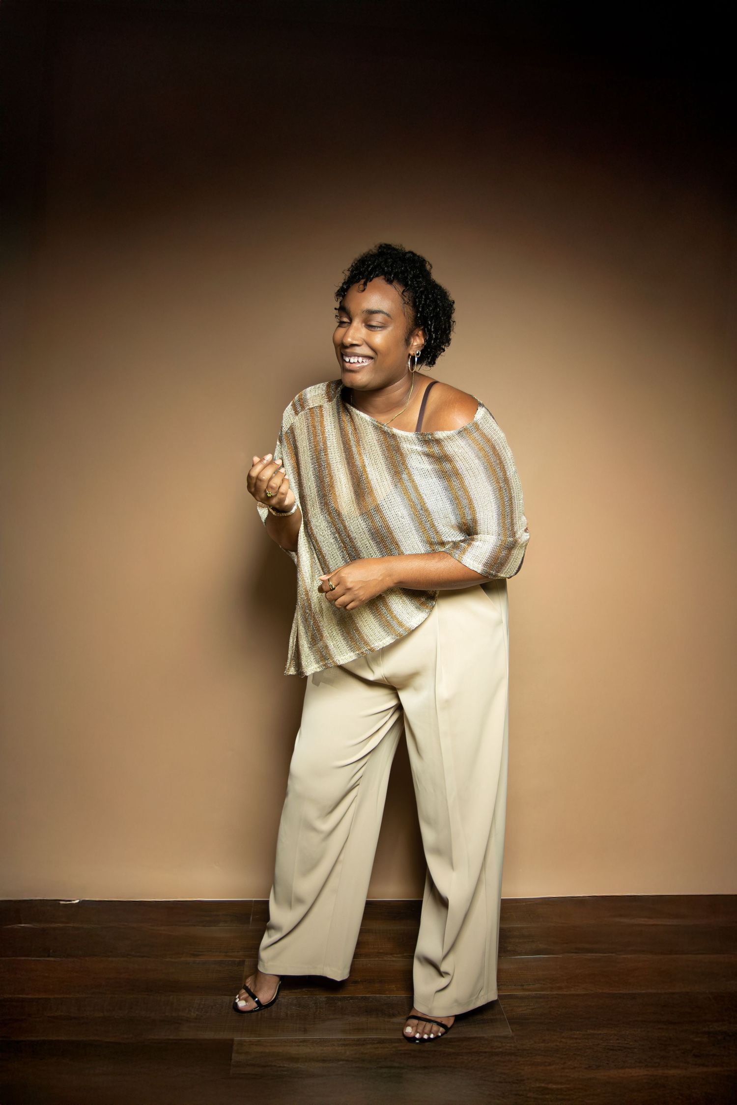 A smiling Black woman poses in cream wide-leg pants and striped top against a warm brown background.