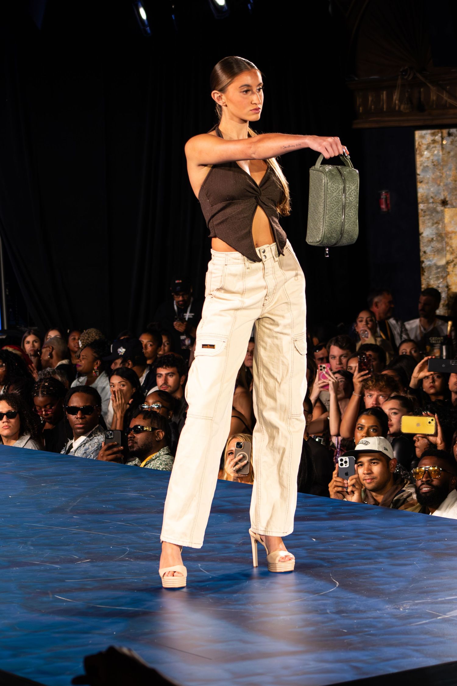Female model walks runway in brown crop top, white cargo pants, and heels carrying green bag.