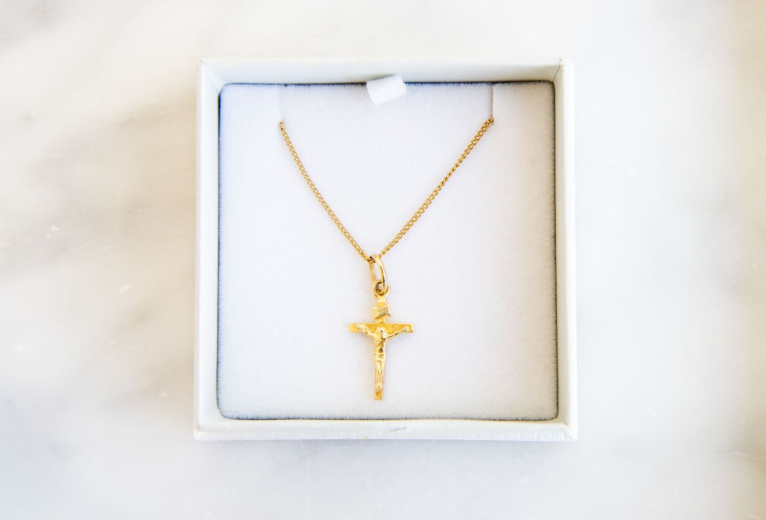 Gold crucifix pendant necklace displayed in a white gift box on a marble surface.