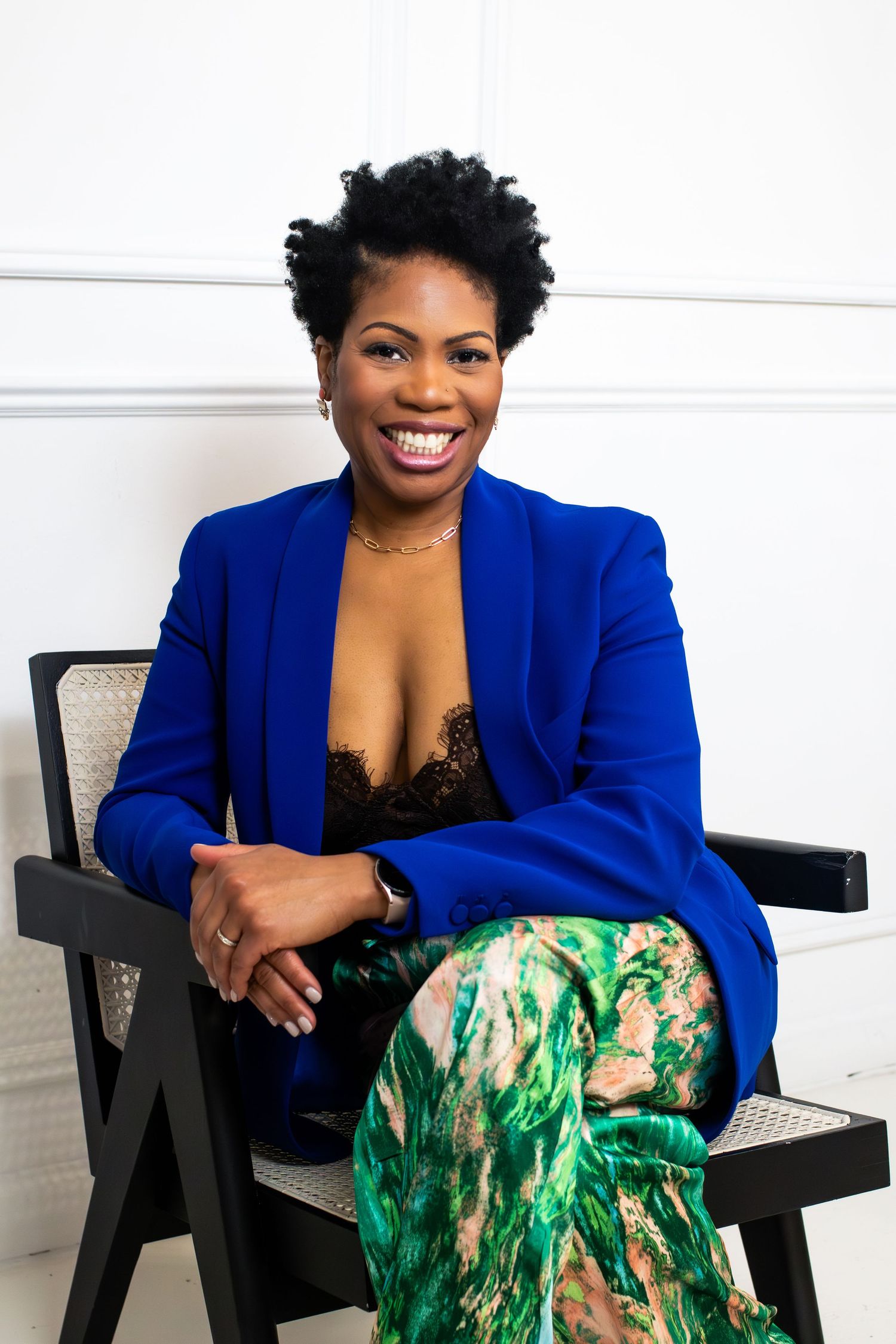 Professional portrait of person in royal blue blazer and patterned green pants seated in modern chair.