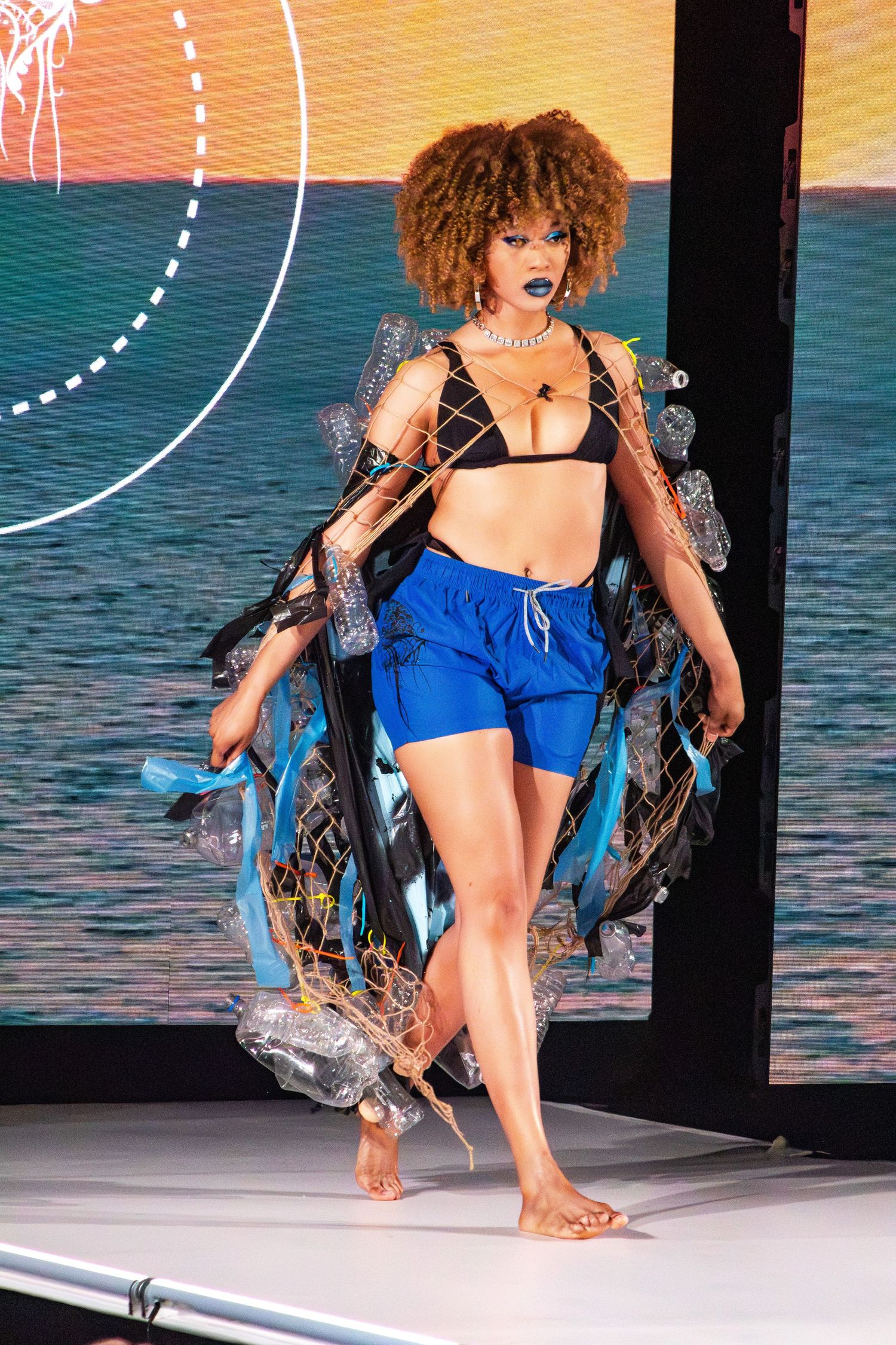 Model walks barefoot on catwalk in blue shorts and sheer cover-up against beach scene.