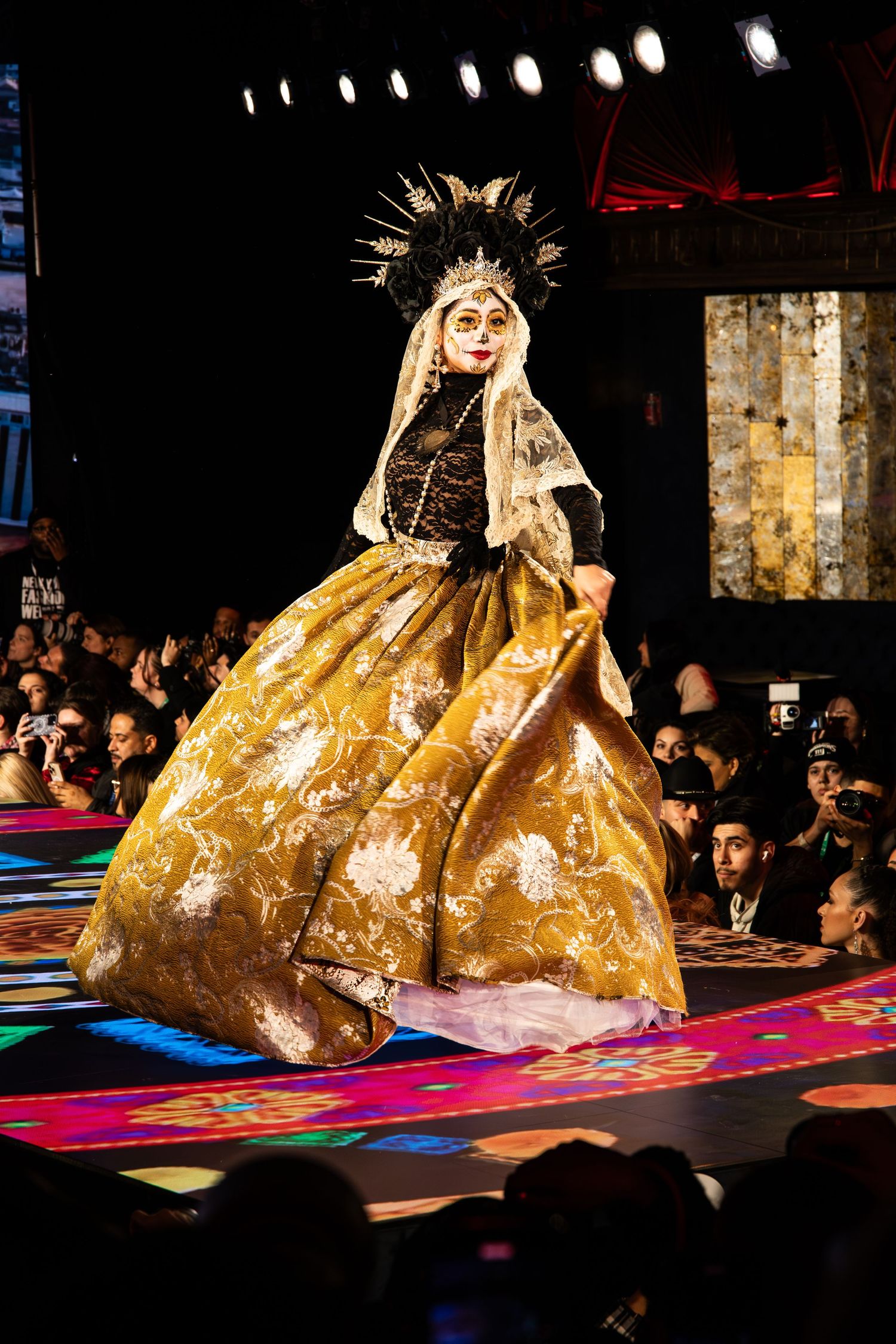 A model walks down a colorful runway wearing an ornate gold ballgown and dramatic crown-like headpiece at a fashion show.