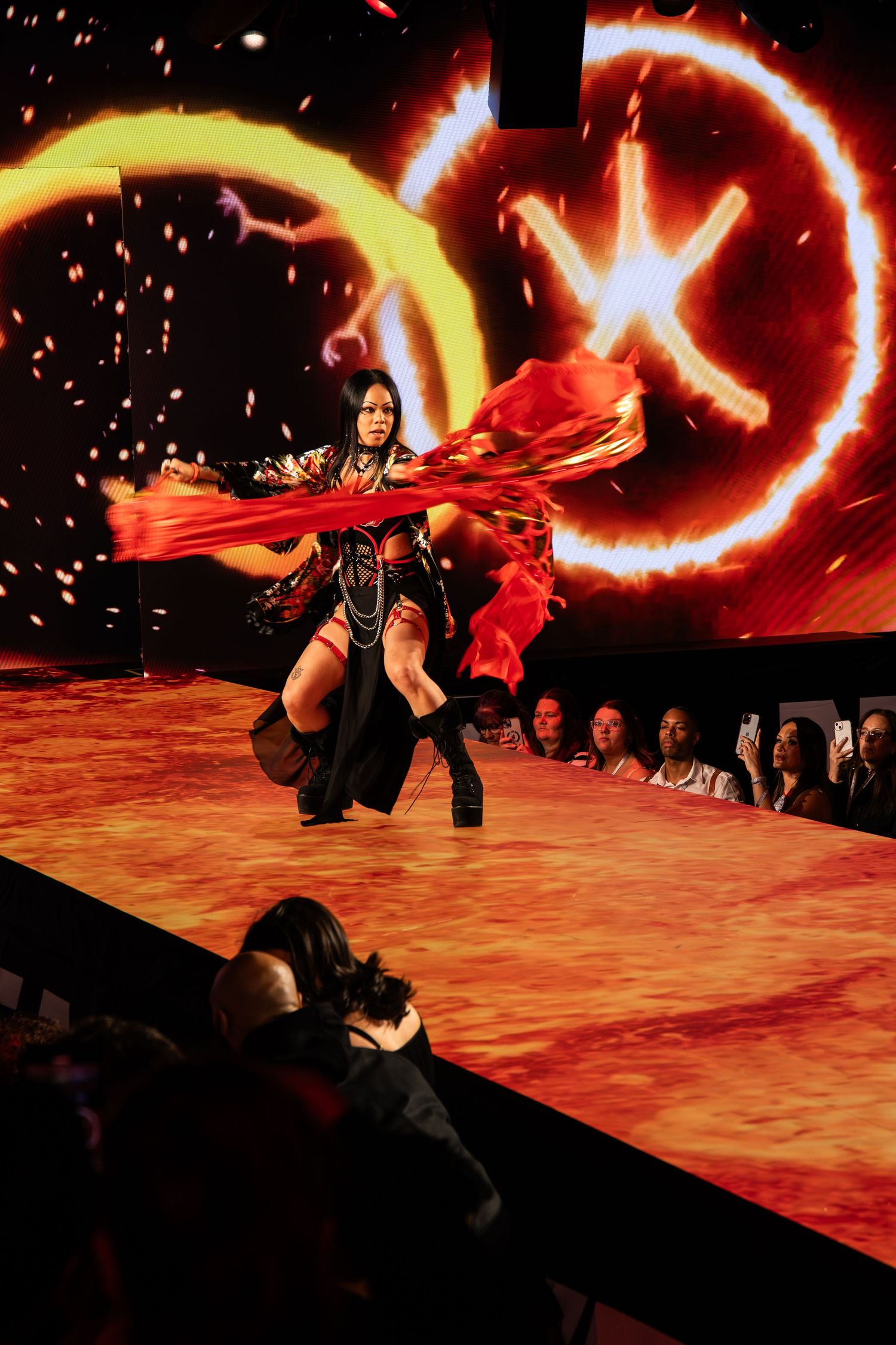 A dancer performs on a fashion runway with dramatic red fabric swirling against glowing circular light effects in the background.