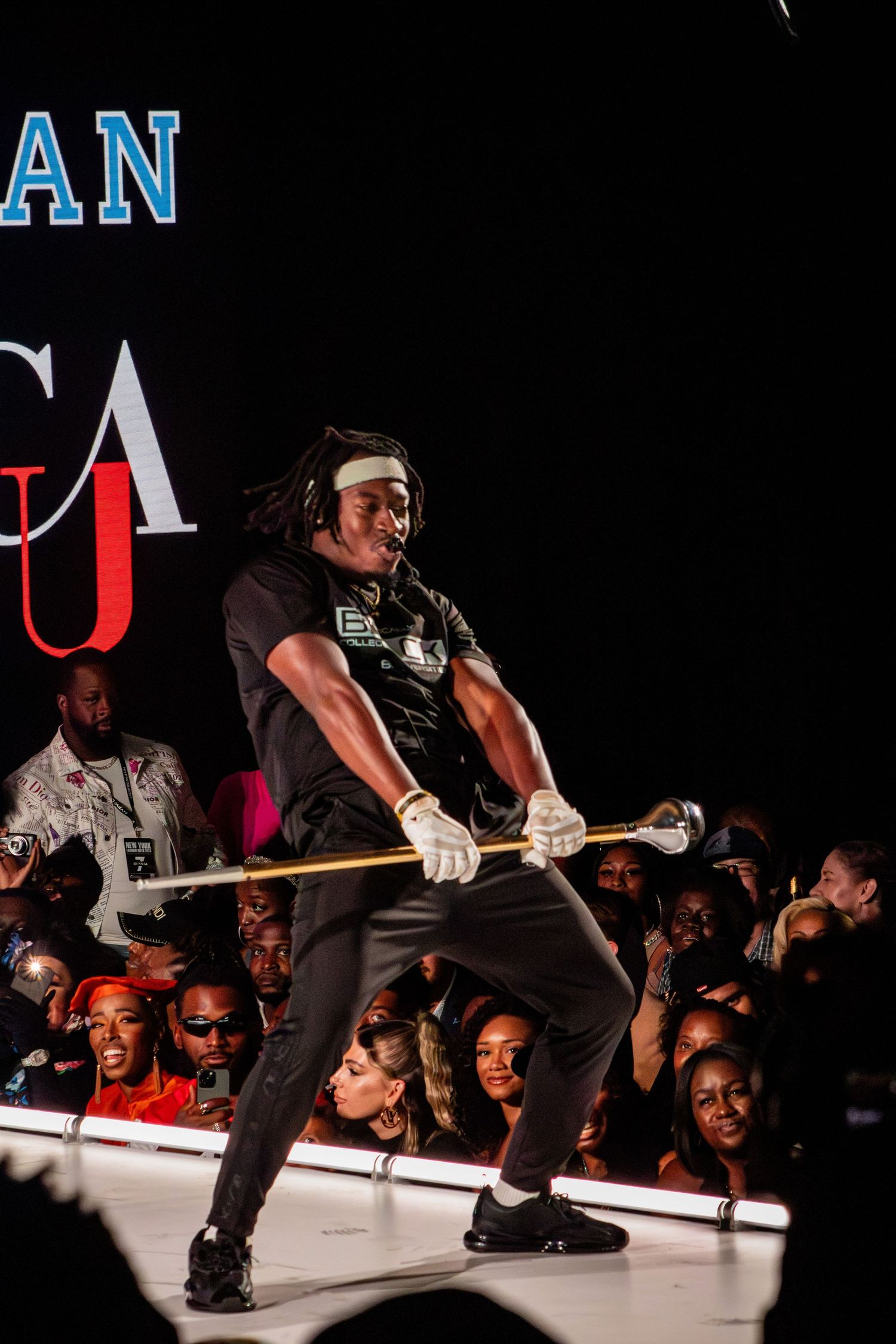 Performer in black outfit jumps while holding a staff on illuminated stage.