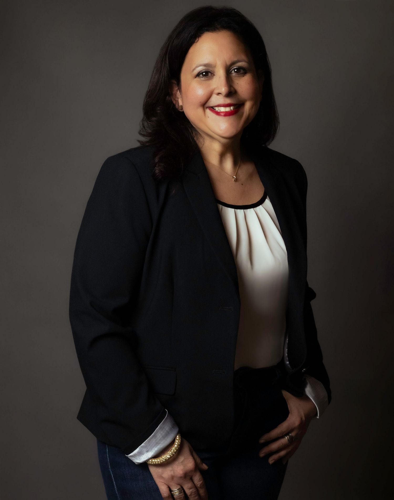 Professional portrait of someone in black blazer and white pleated blouse against dark background.