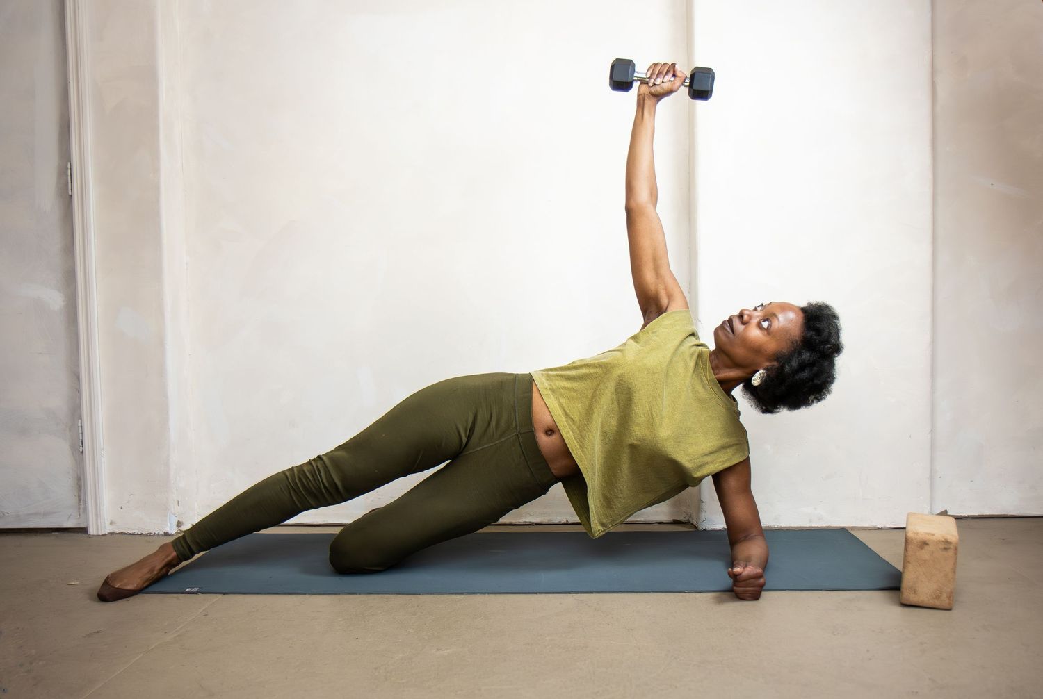 Person performs a side plank exercise on a yoga mat while holding a dumbbell overhead in an olive green outfit.