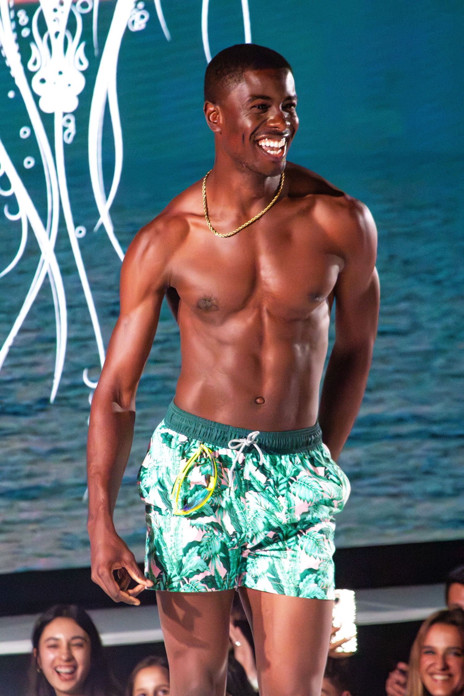 Athletic model showcases green floral swim shorts on beach-themed runway with audience visible.
