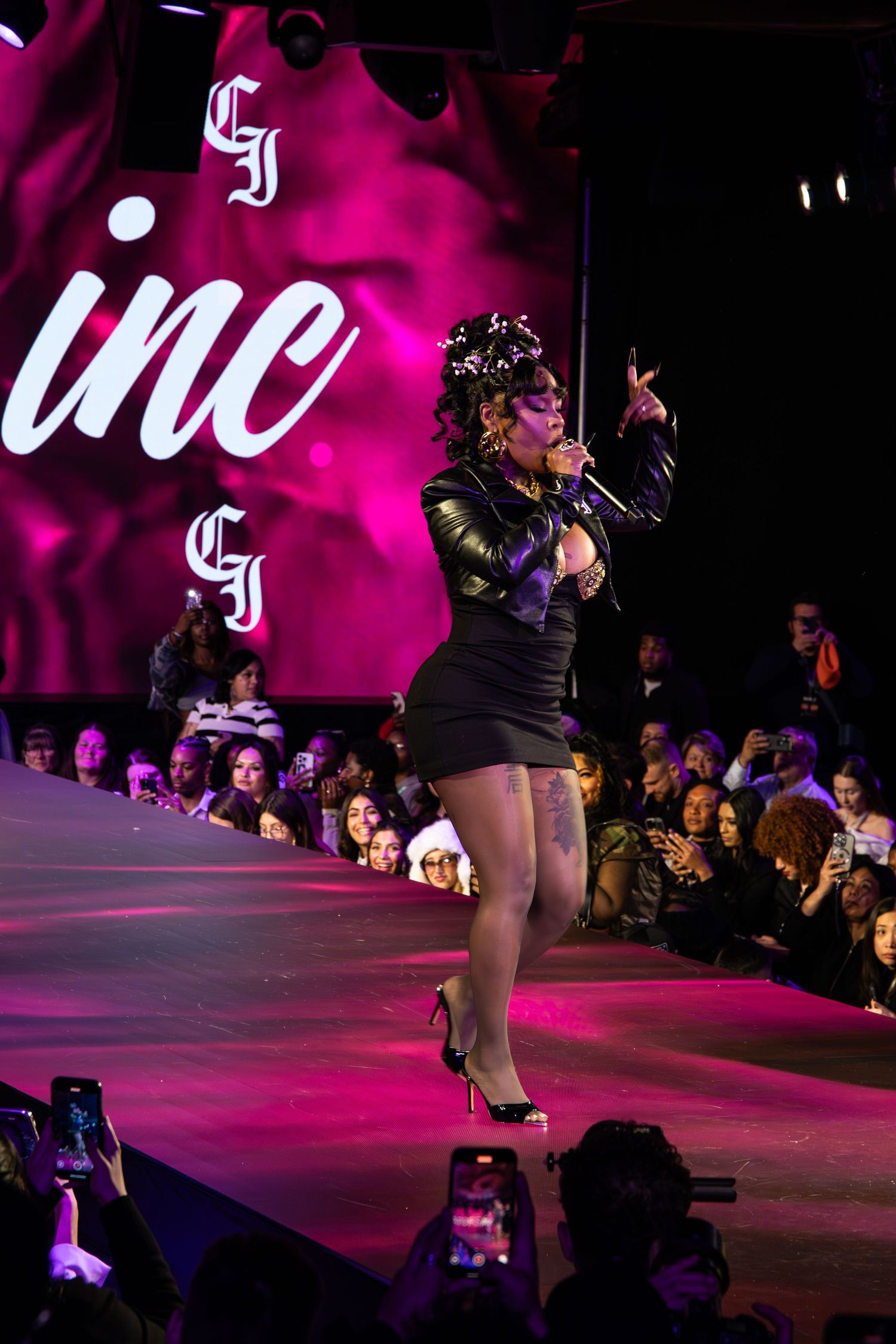 Performer in black dress and crown strikes a pose on pink-lit runway stage during fashion show performance.