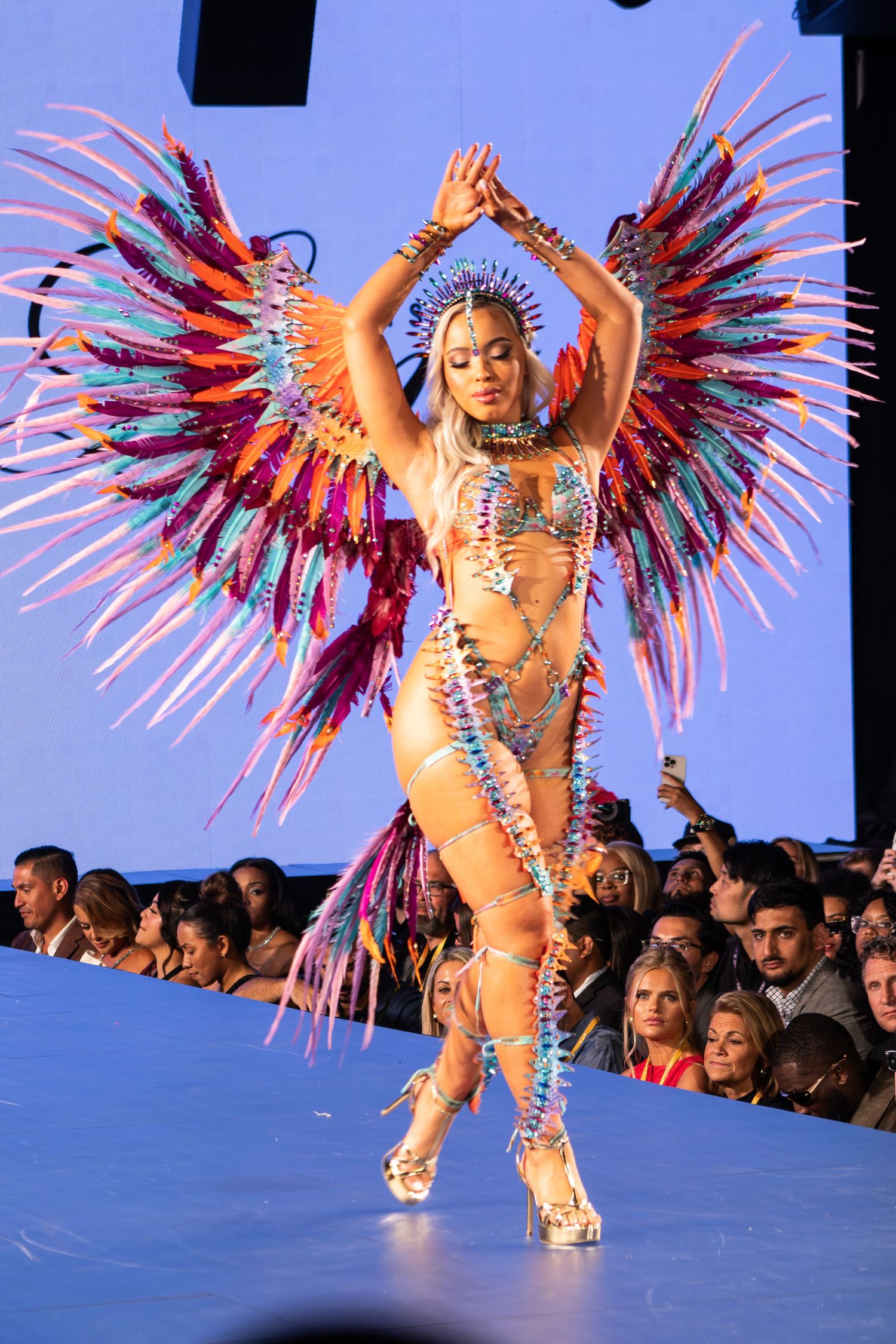 Model poses on runway in colorful feathered wings bikini costume with crystal headpiece.