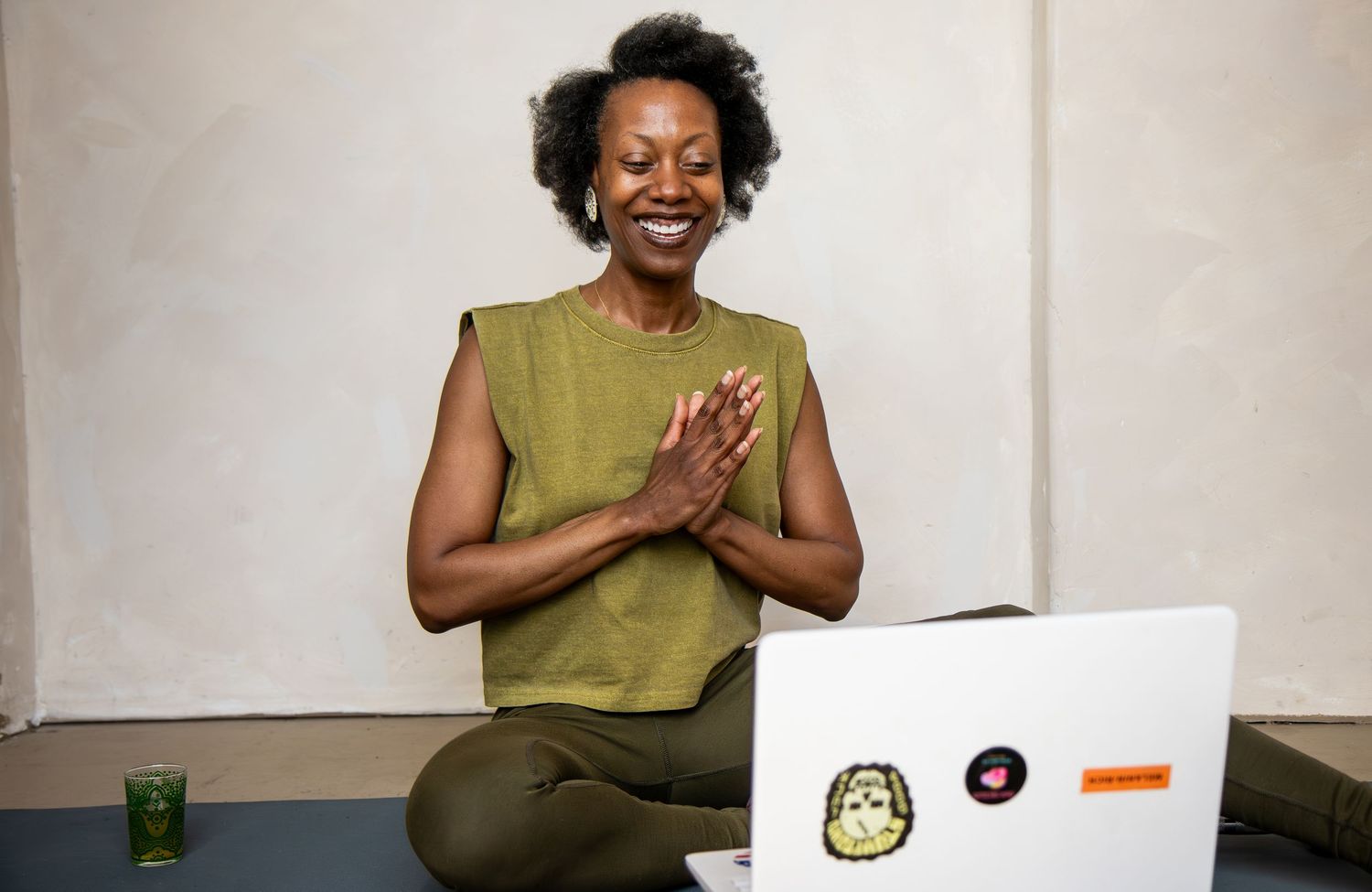 A person in olive green attire sits at a desk with a laptop, smiling warmly with hands clasped together.