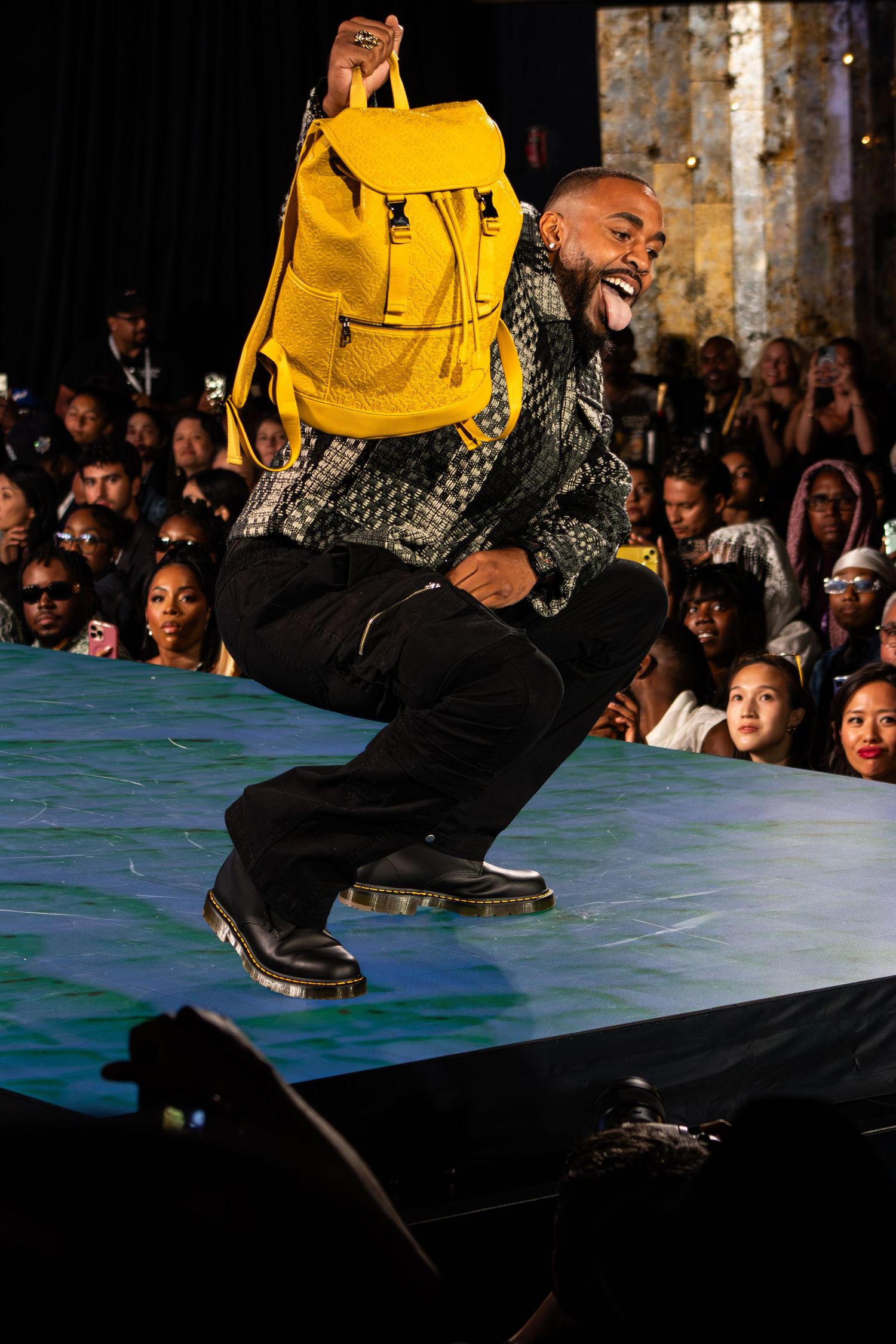 Man jumping on runway holding yellow backpack at fashion show.