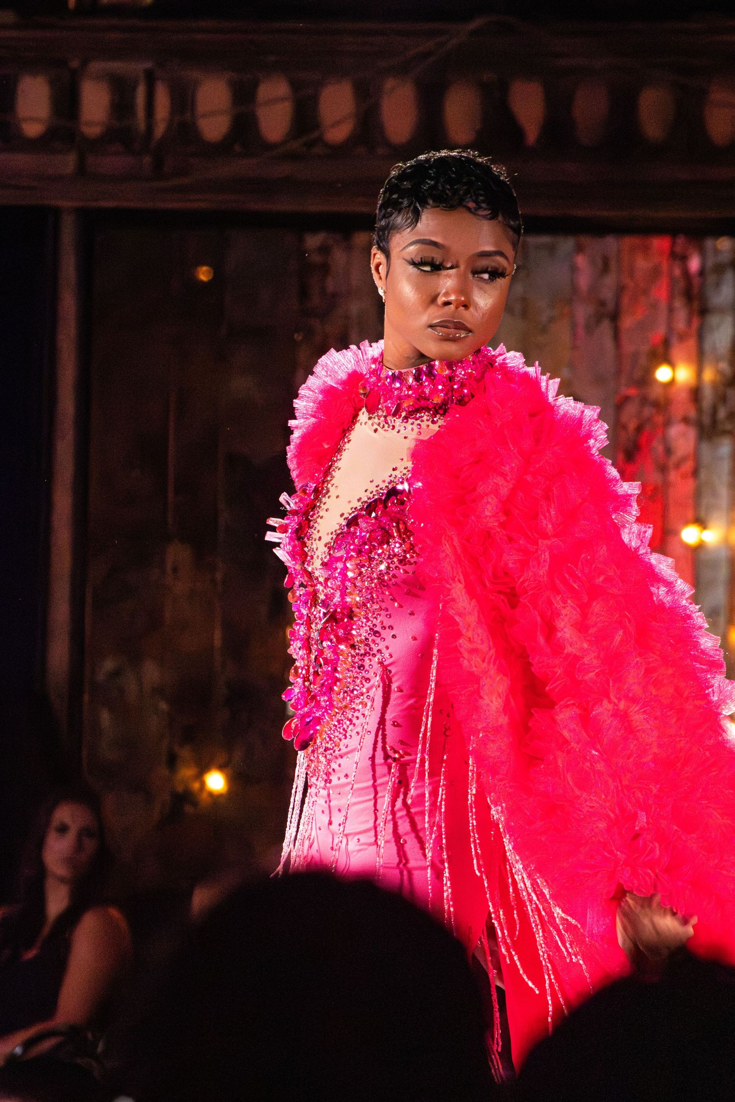 Fashion model in vibrant hot pink feathered dress and sequins performing on dimly lit stage.