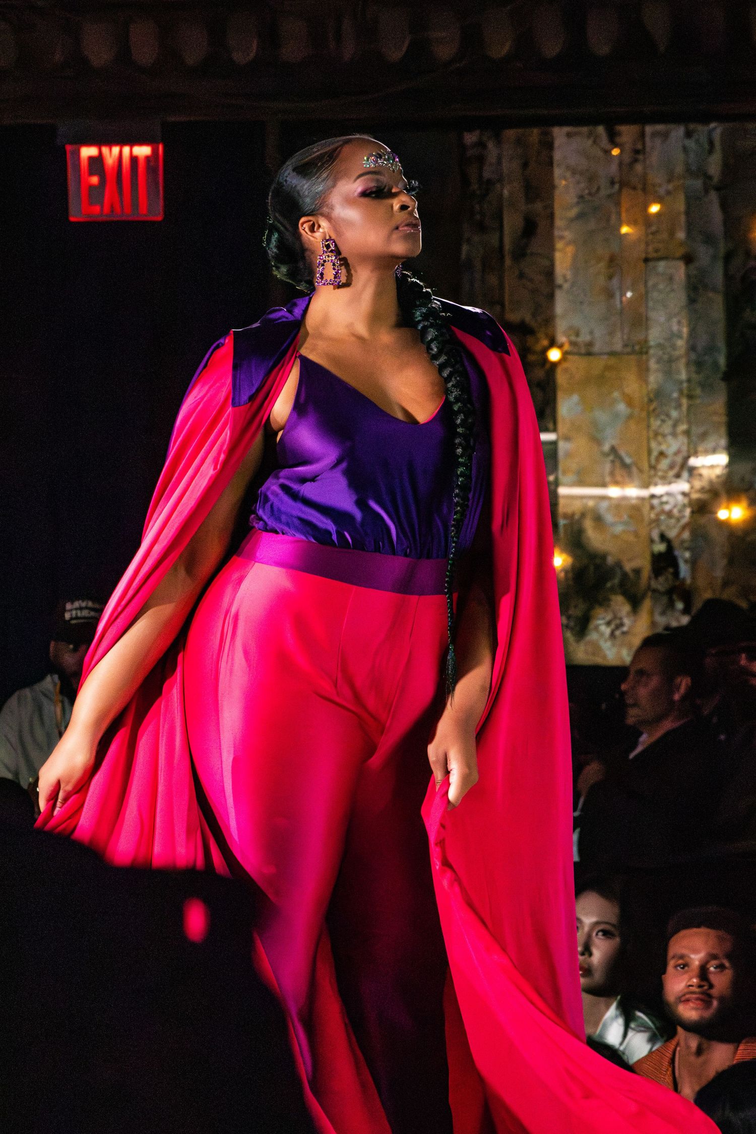Fashion model walks runway in dramatic pink and purple evening gown with flowing cape during fashion show.
